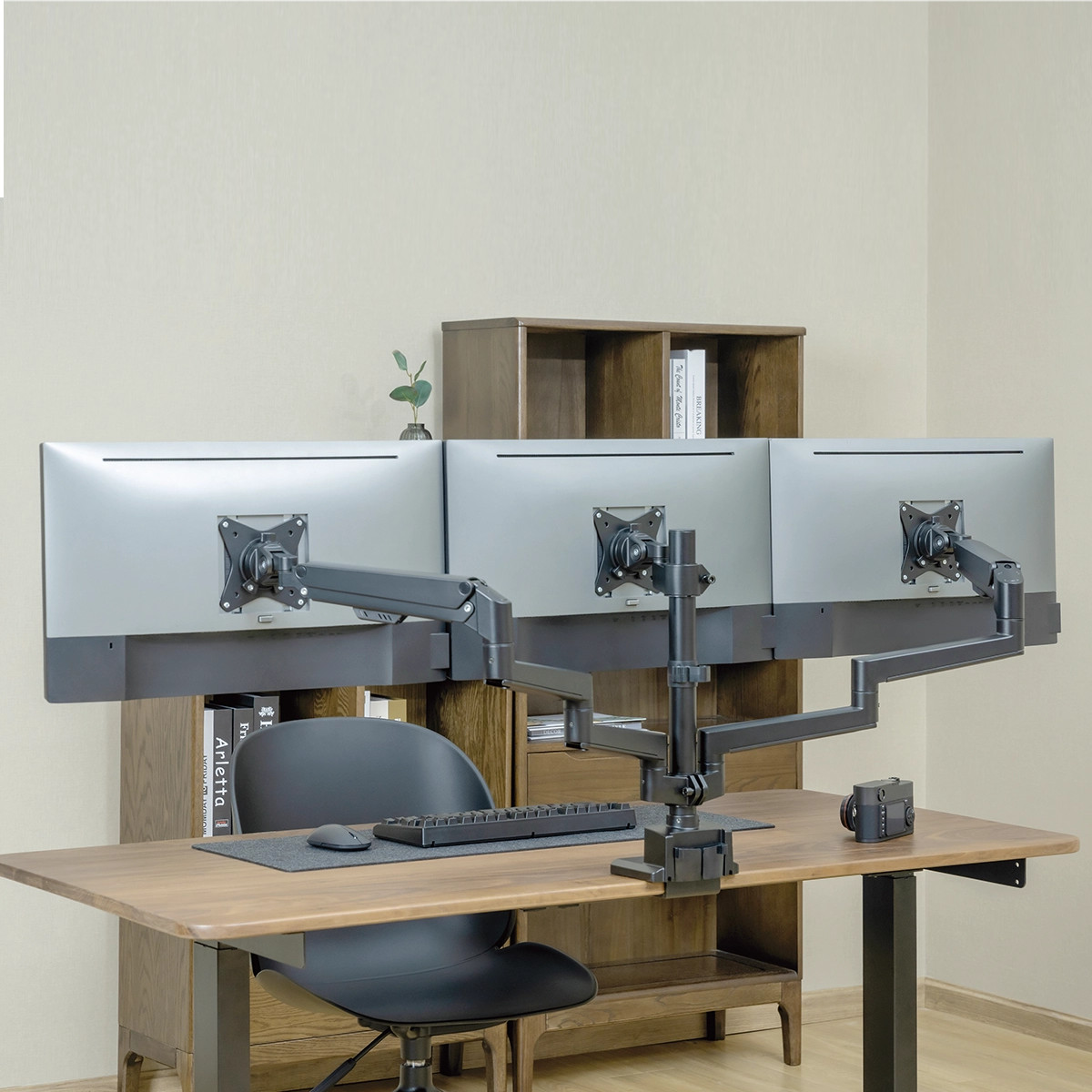 Drei Monitore an schwarzen Armen, die an einem Schreibtisch befestigt sind. Hintergrund ist ein helles Zimmer.