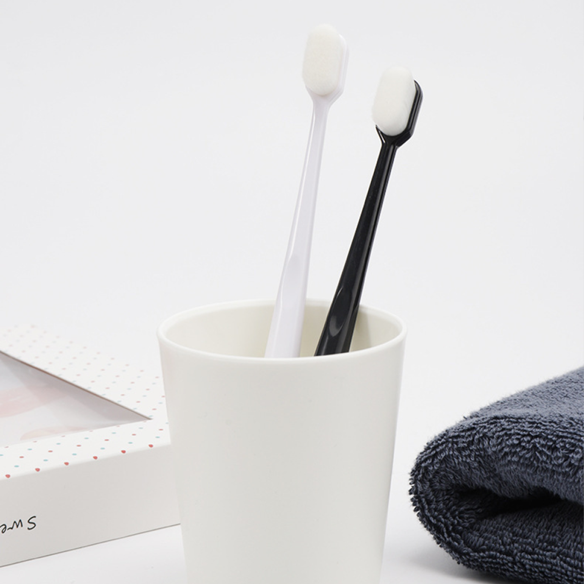 Zwei Zahnbürsten in einem weißen Becher, Handtuch und Geschenkbox, alles vor weißem Hintergrund.