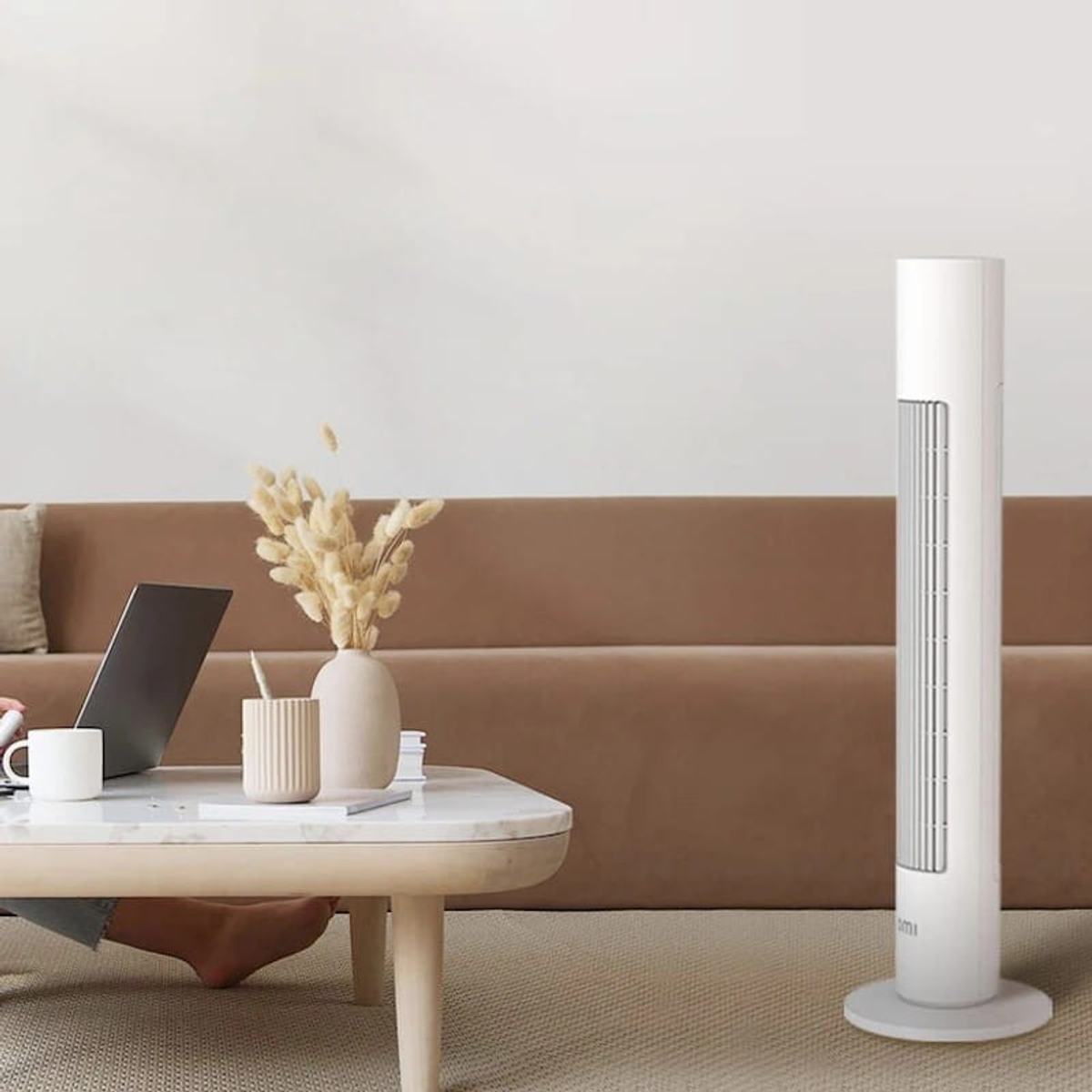 Un ventilatore a torre bianco in un soggiorno con divano marrone, tavolo e laptop. Tappeto beige chiaro.