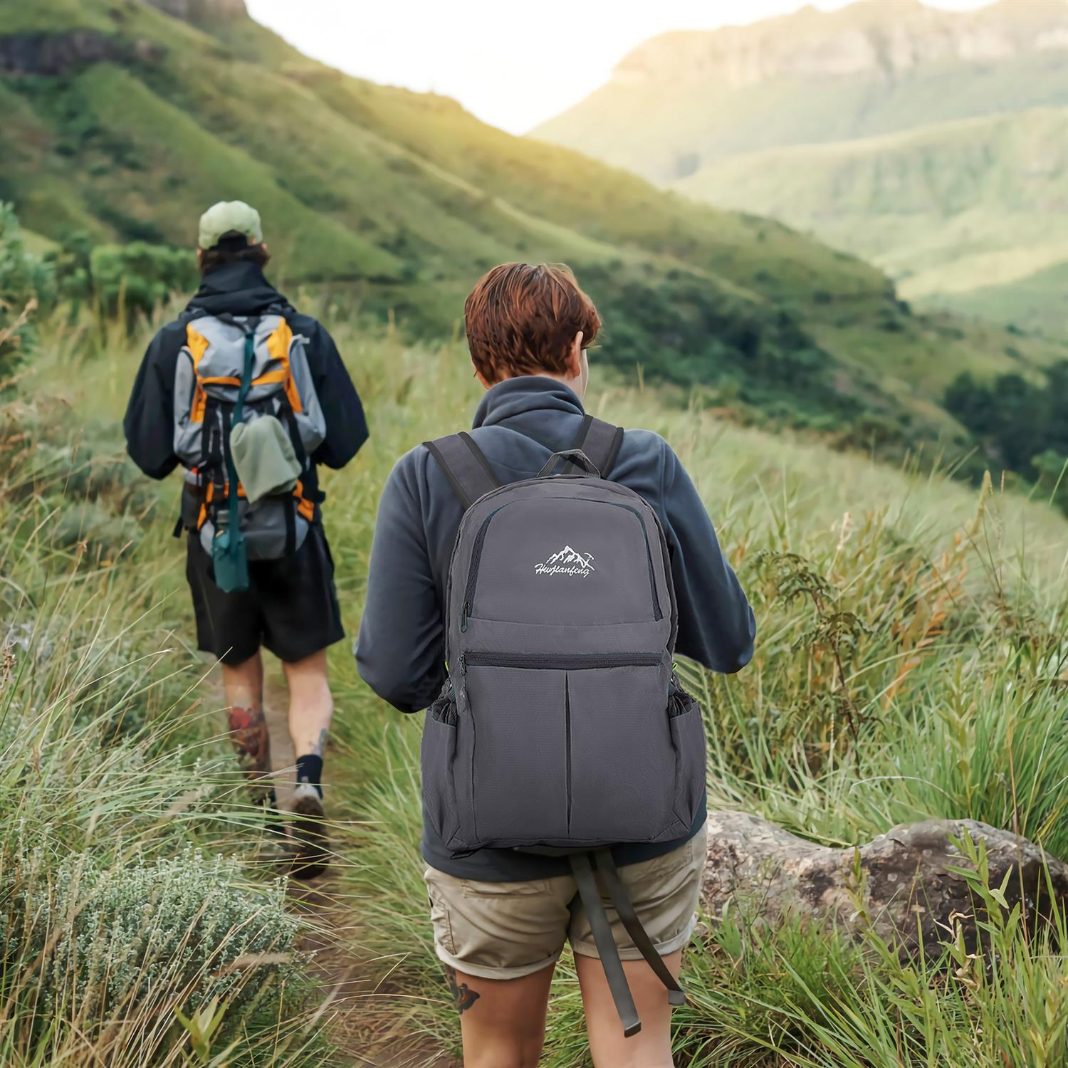 Zwei Wanderer mit Rucksäcken gehen auf einem Pfad durch eine grüne Berglandschaft.