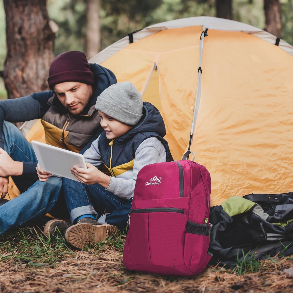 Vater und Sohn schauen vor einem Zelt auf ein Tablet, ein Rucksack ist in der Nähe.