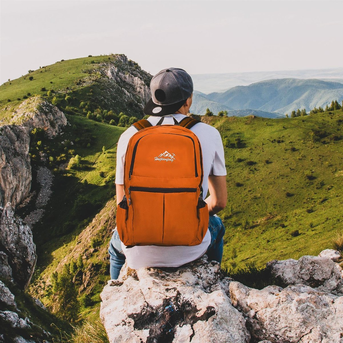 Person mit orangefarbenem Rucksack sitzt auf Felsen und blickt auf Berglandschaft.