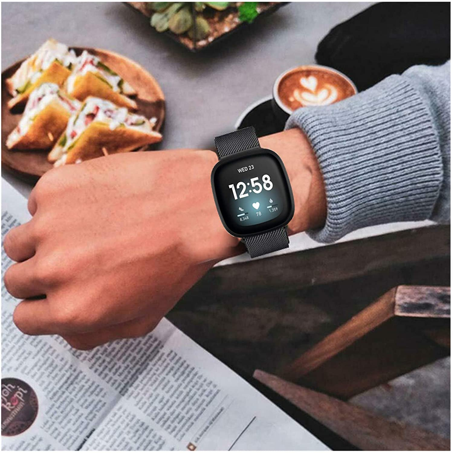 Handgelenk mit schwarzer Smartwatch, die Zeit, Herzfrequenz und Schritte anzeigt. Sandwiches und Kaffee in der Nähe.