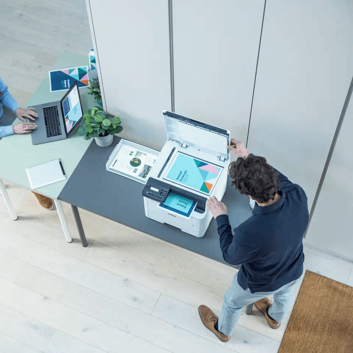 Mężczyzna otwierający drukarkę, a w tle dwie osoby pracują na laptopie i tablecie.