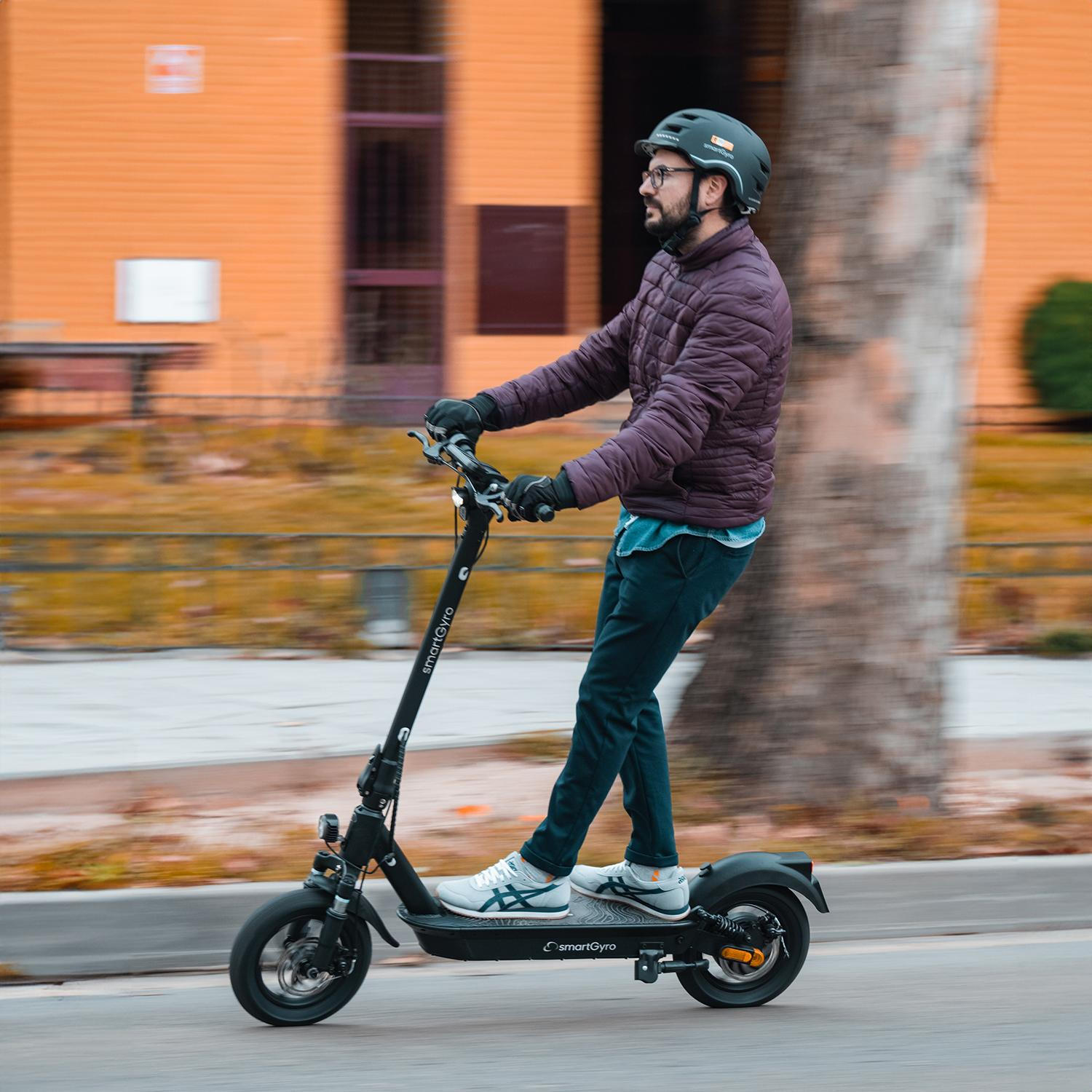 Mężczyzna na czarnym skuterze, w kasku, jadący drogą. Pomarańczowy budynek w tle.