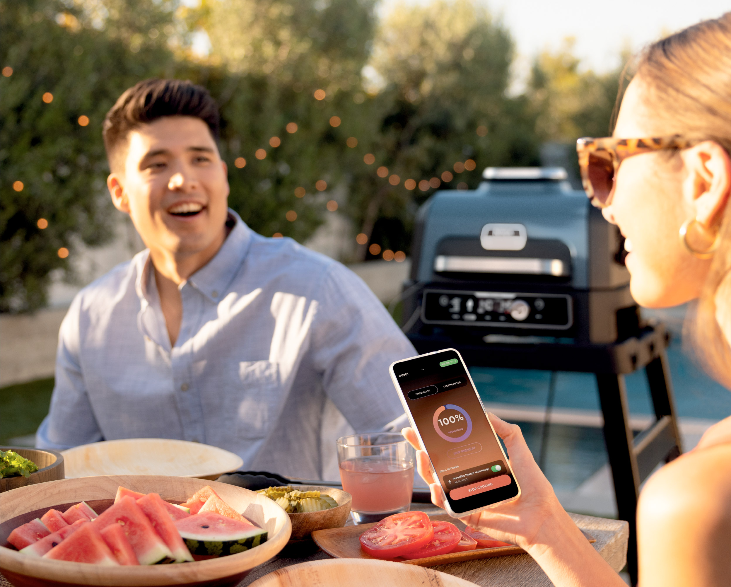 Man en vrouw. Vrouw houdt telefoon met app vast. Grill en eten op tafel.