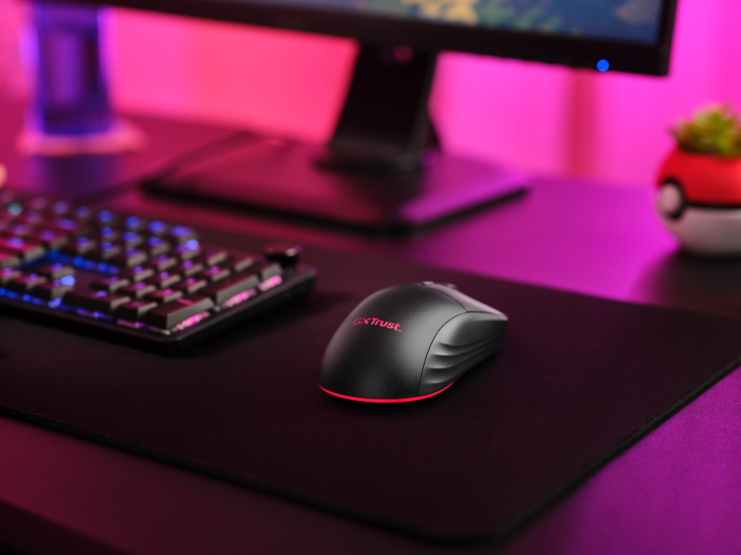 Czarna mysz komputerowa na czarnej podkładce z klawiaturą i monitorem w tle.