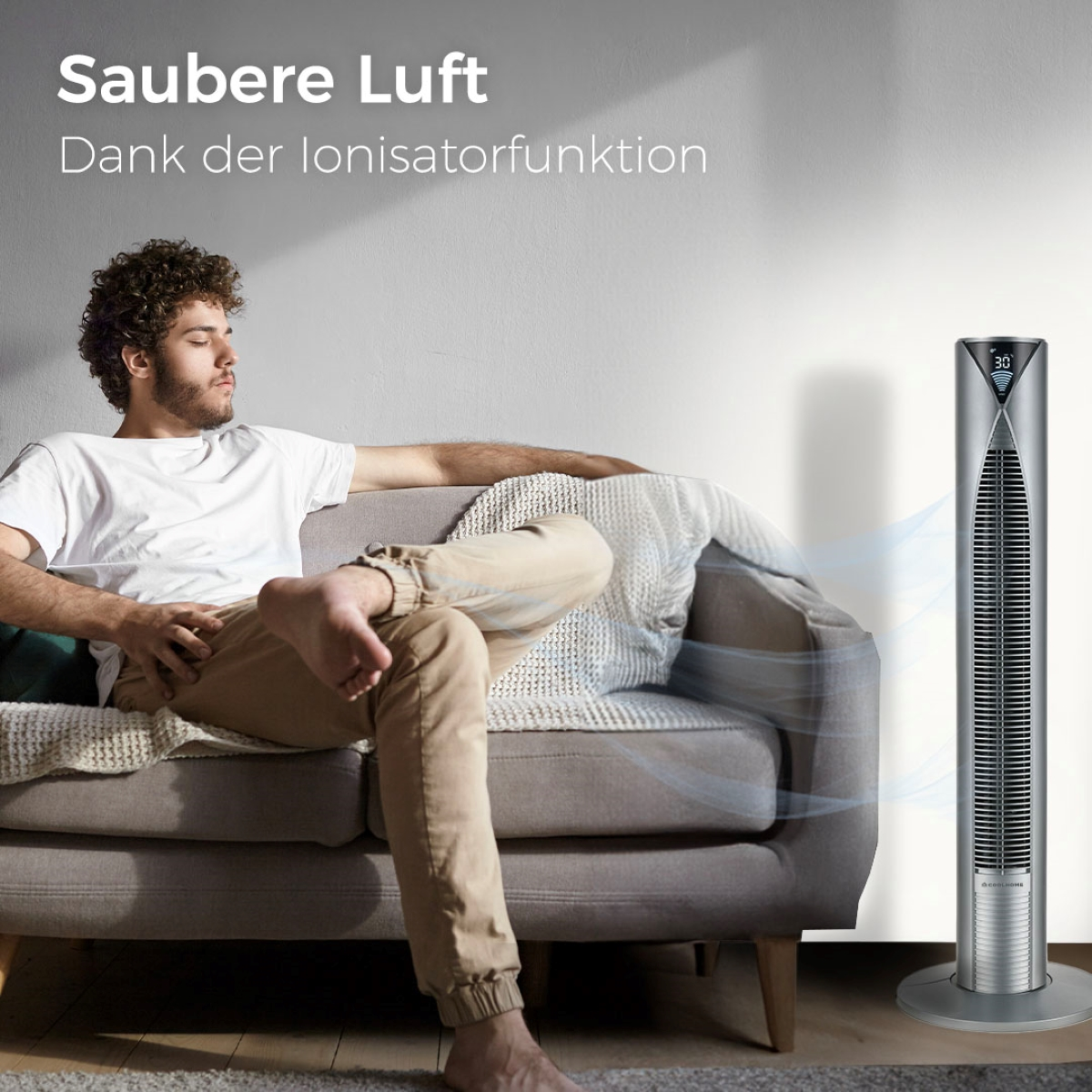Ein Mann sitzt auf einem Sofa, im Hintergrund steht ein hoher silberner Ventilator, der Luft bläst.