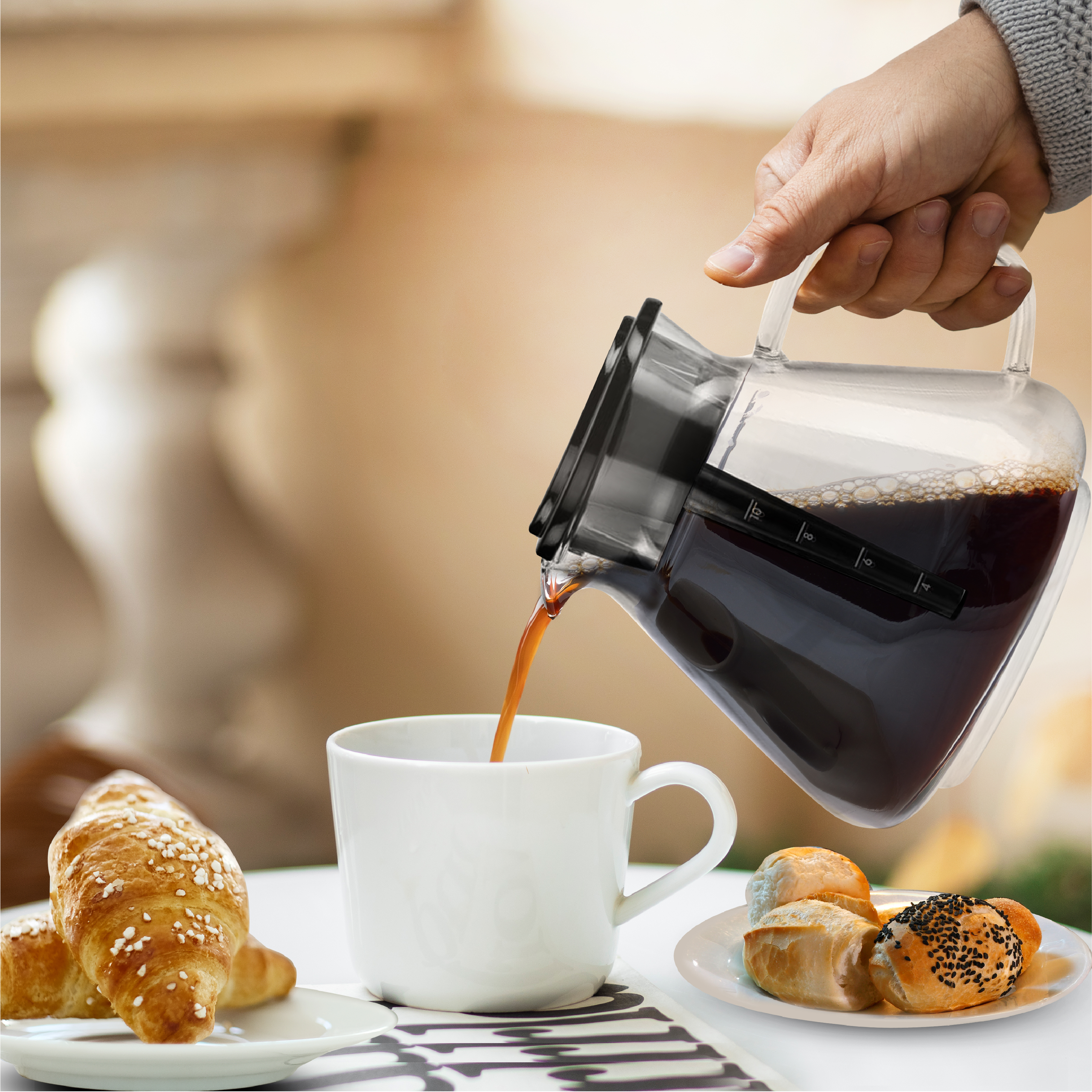 Koffie wordt uit een glazen kan in een witte mok geschonken, met gebak op een tafel.
