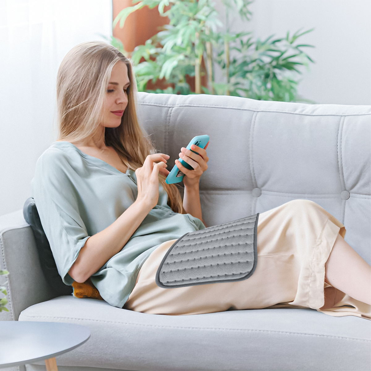 Woman on a sofa using a phone, with a cushion and a heating pad on her lap.