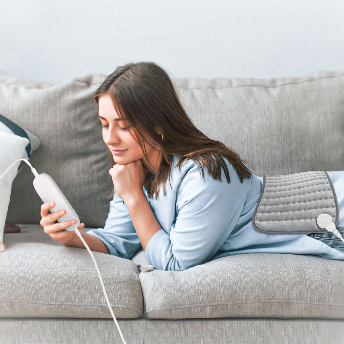 Woman on a sofa with a heating pad on her back, holding a control. The background is a wall.