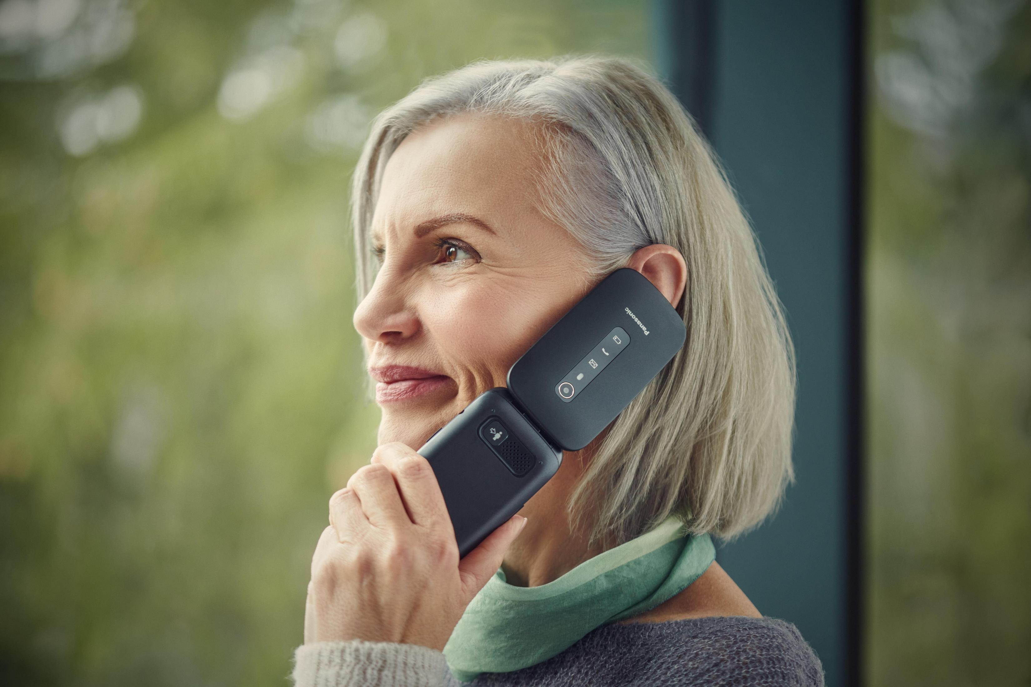 Woman holding a black flip phone. She is smiling and looking to the right. Green scarf is around her neck.