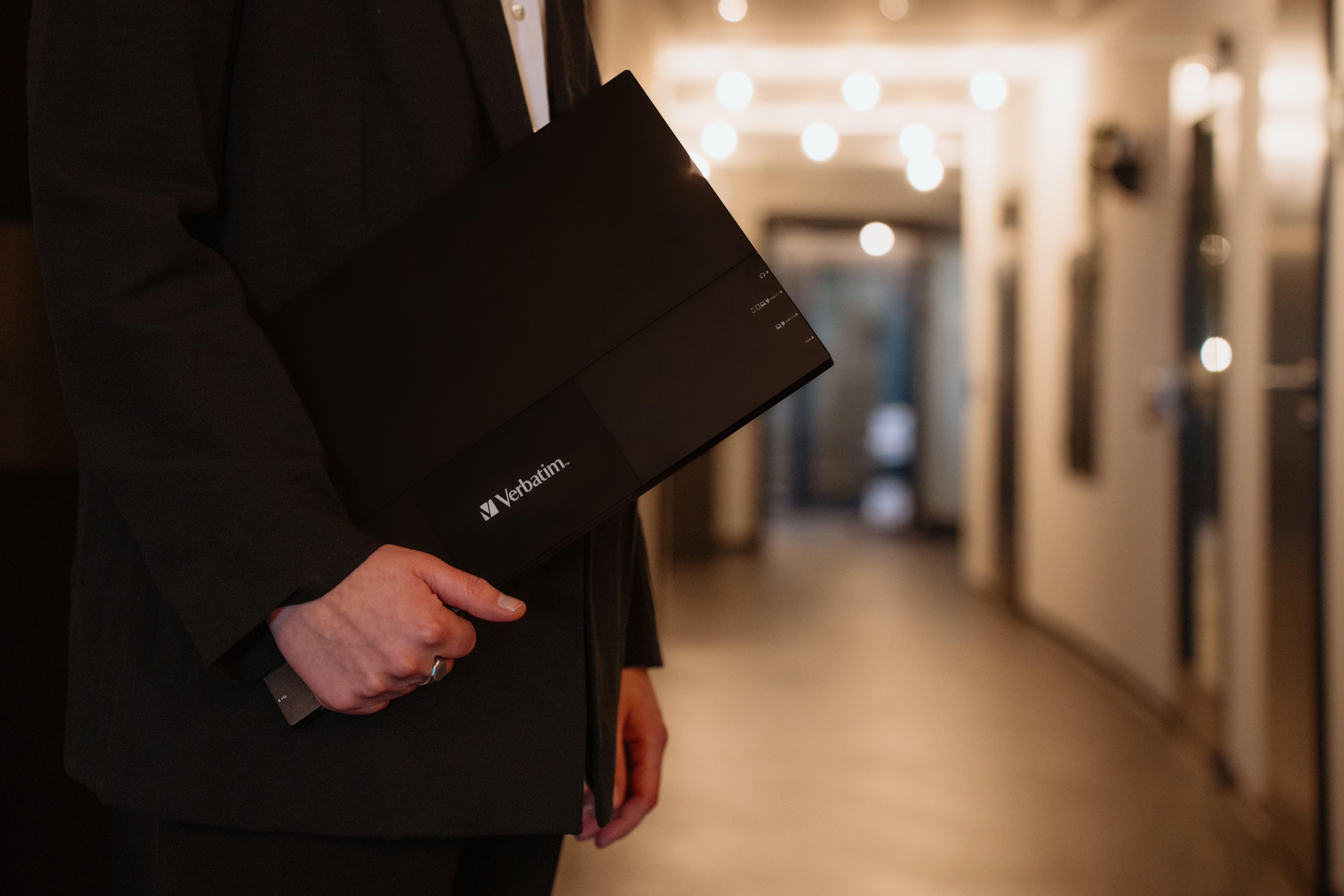 Osoba w garniturze trzymająca czarny folder Verbatim, idąca korytarzem, rozmyte tło.