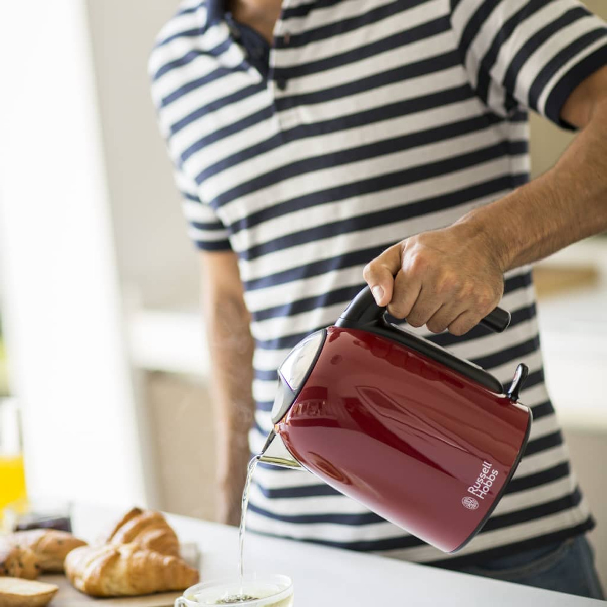 Mężczyzna nalewa wodę z czerwonego czajnika do szklanki, rogaliki na pierwszym planie.
