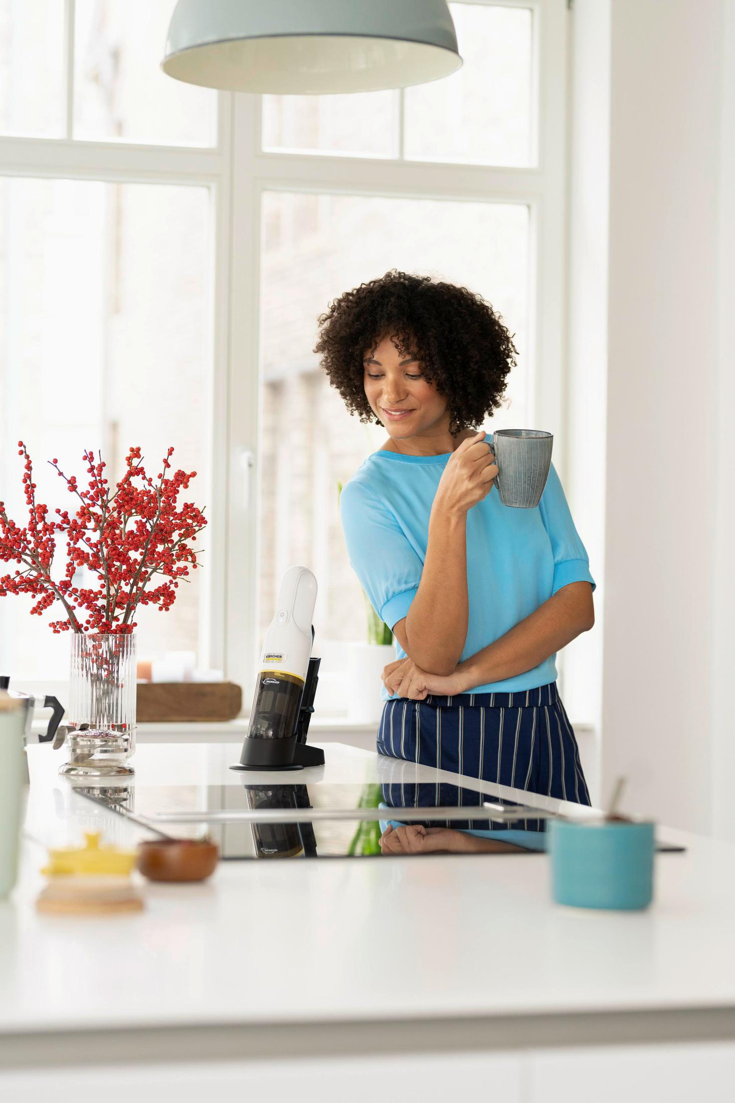 Femme en haut bleu tenant une tasse, appuyée sur un comptoir dans une cuisine avec des fleurs.