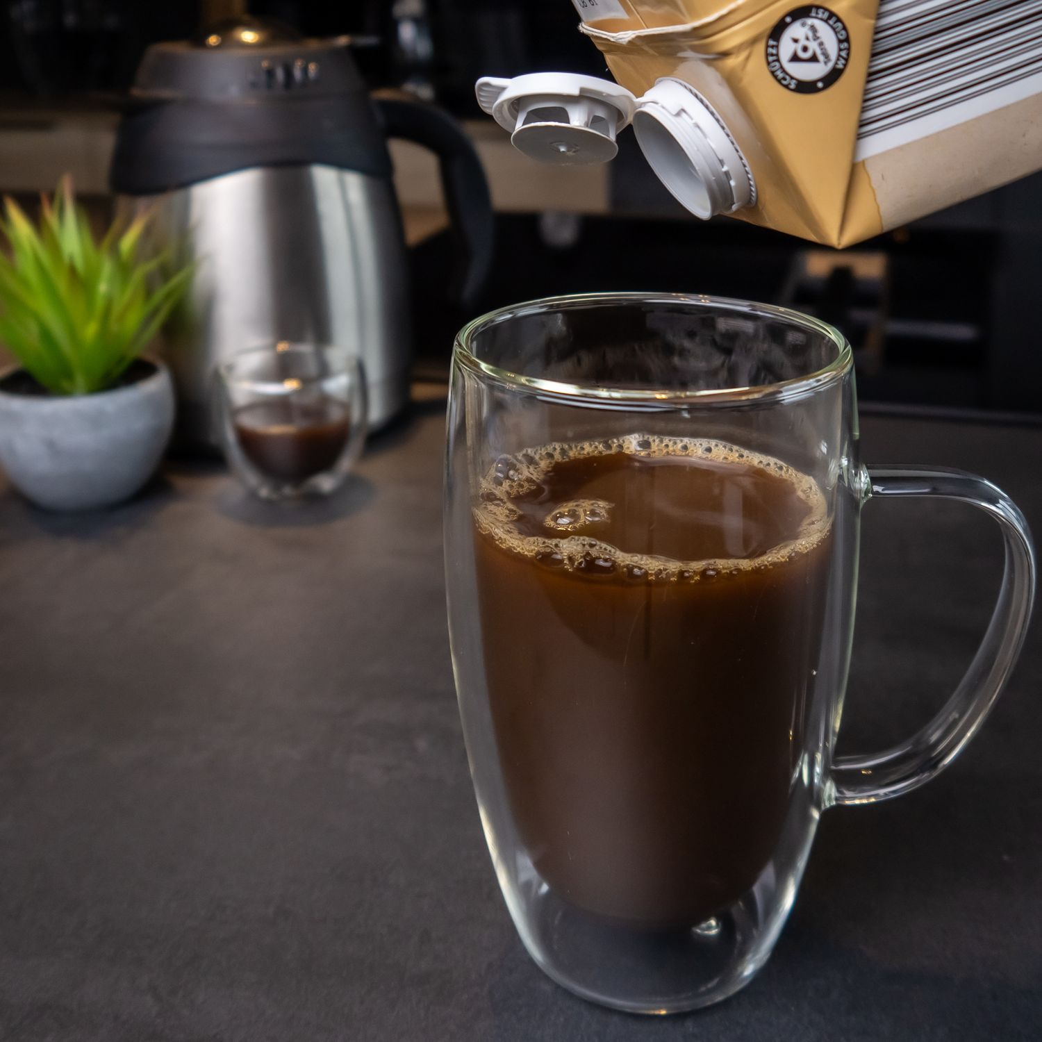Kaffee in einem Glas. Milch wird aus einem Karton gegossen. Schwarze Arbeitsplatte und Hintergrund.