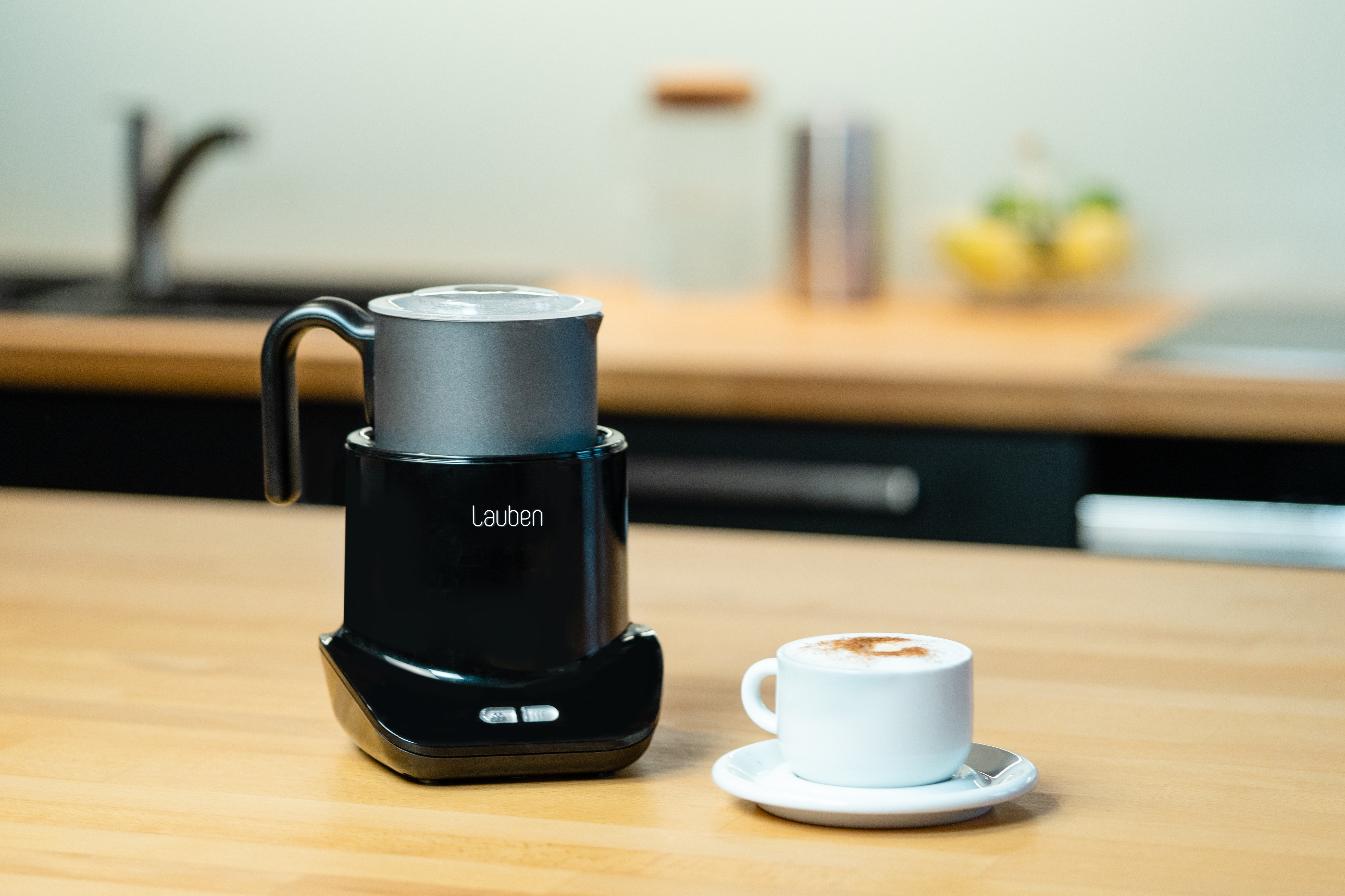 Ein schwarzer und silberner Milchaufschäumer auf Holz. Rechts steht eine Tasse Kaffee.
