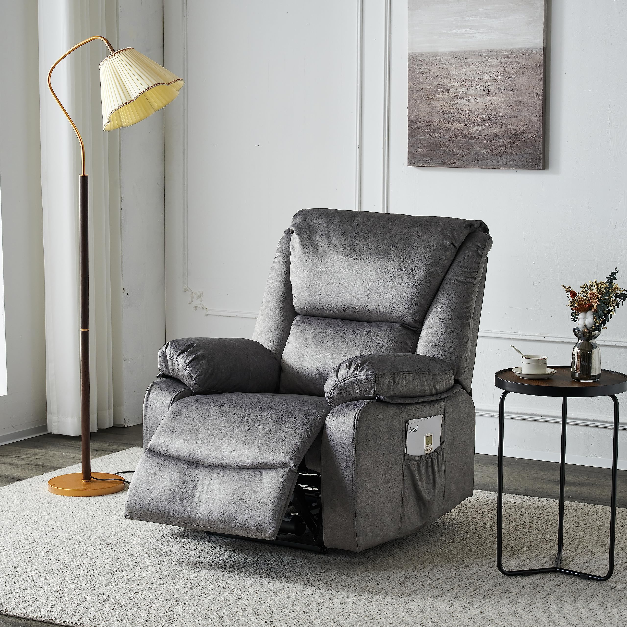 Sillón reclinable gris en un salón, con lámpara, mesa y obras de arte en el fondo.