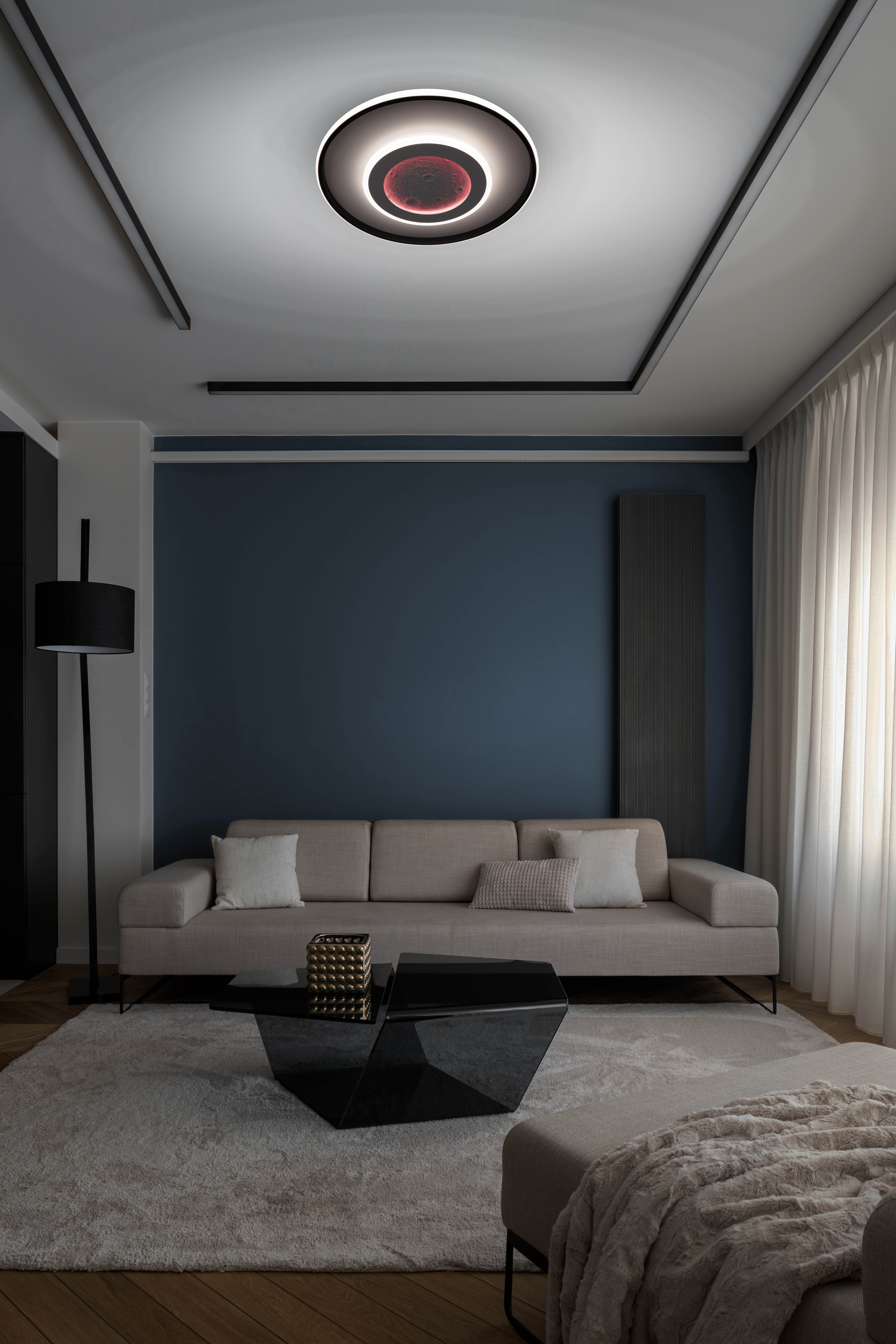 Innenaufnahme eines Wohnzimmers. Ein beiges Sofa, ein schwarzer Couchtisch und eine große Deckenleuchte.