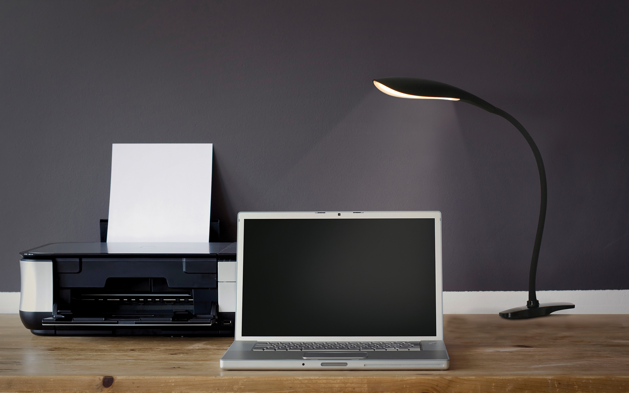 Laptop, Drucker und Schreibtischlampe auf einem Holzschreibtisch vor einer dunkelgrauen Wand. Die Lampe leuchtet.