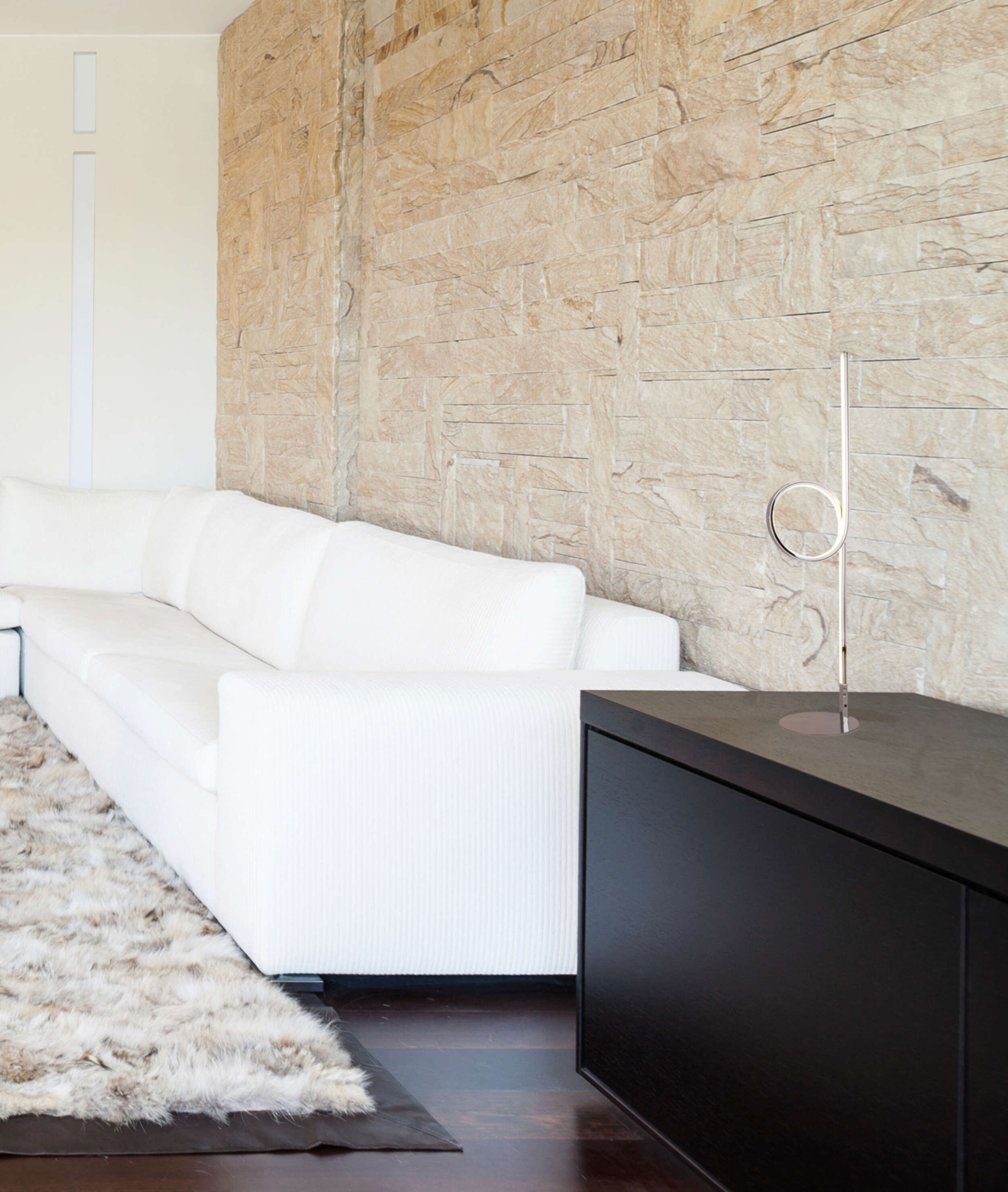 Modernes Wohnzimmer mit weißem Sofa, dekorativer Skulptur und schwarzem Schrank. Beige Steinmauer.