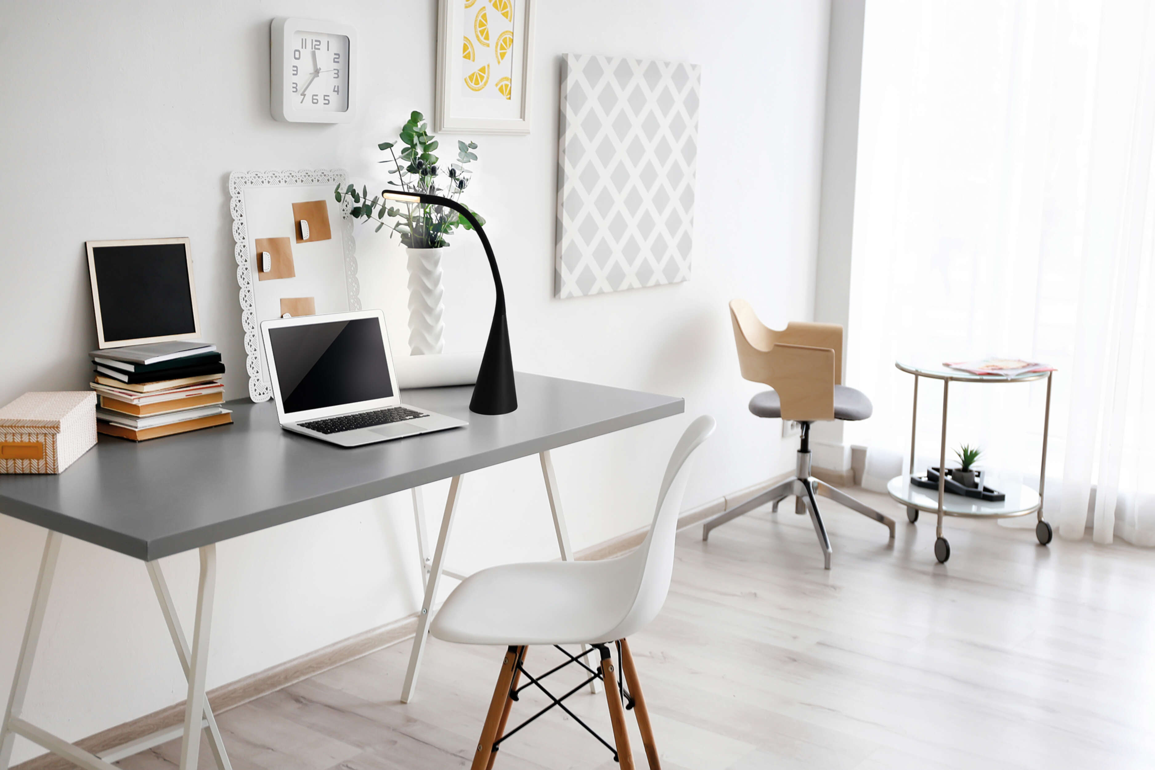Büro mit Schreibtisch, Laptop und schwarzer Lampe. Der Hintergrund ist eine weiße Wand.