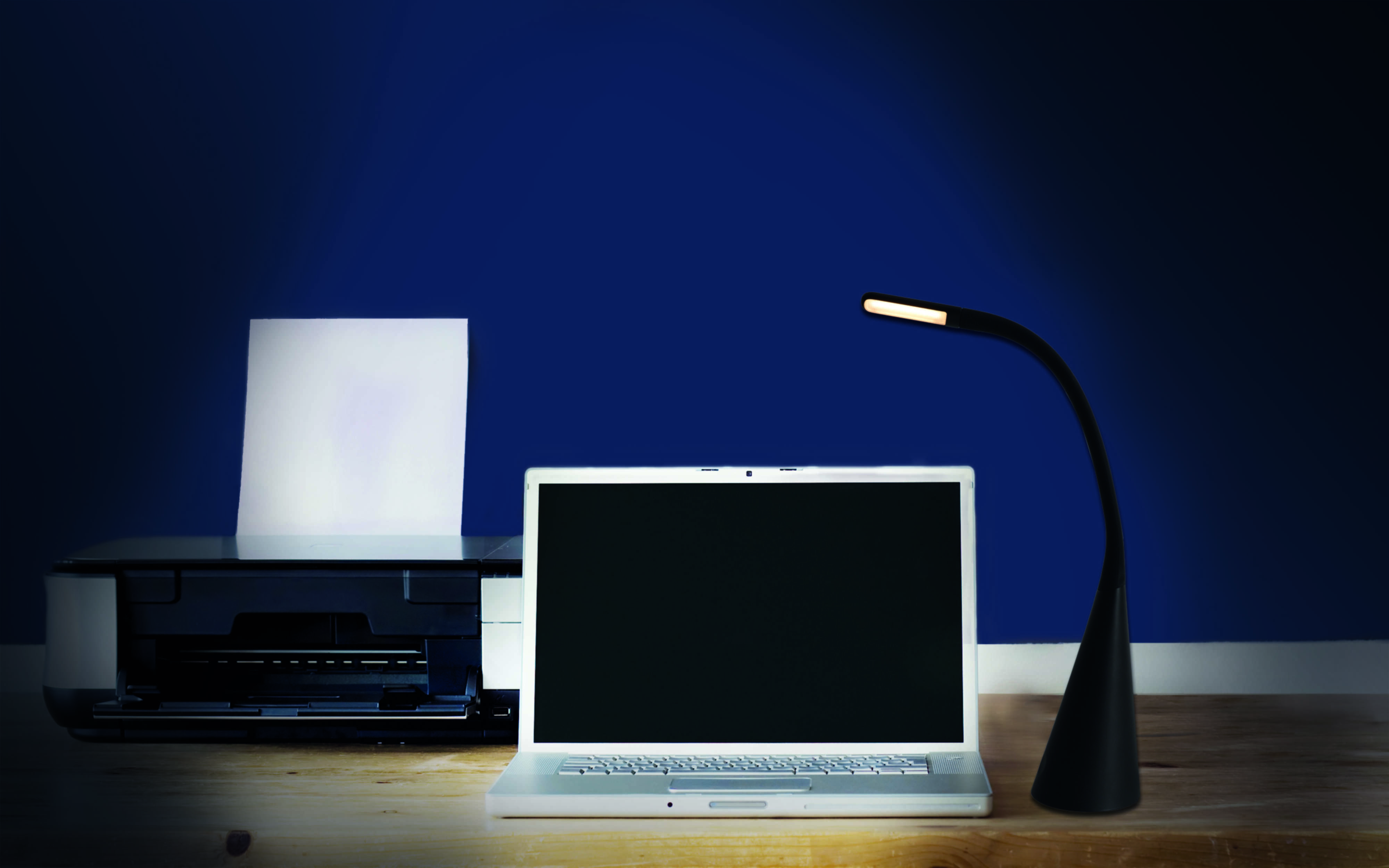 Laptop, Drucker und eine schwarze Schreibtischlampe auf einem Holztisch. Eine blaue Wand im Hintergrund.