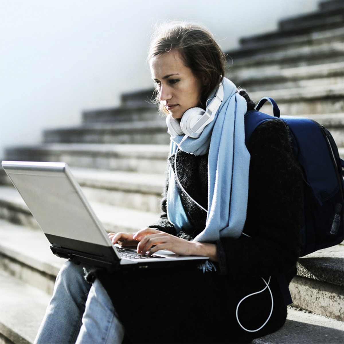 Frau sitzt auf Treppen, benutzt einen Laptop mit Kopfhörern und einem Schal.