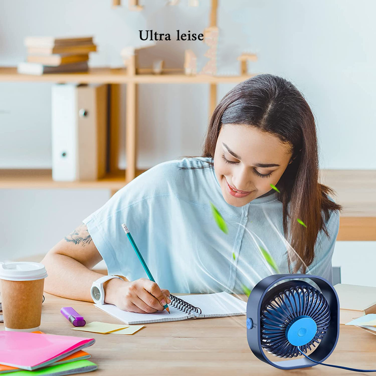 Eine Frau schreibt mit einem Stift in ein Notizbuch, neben einer Tasse, mit einem Ventilator auf dem Schreibtisch.