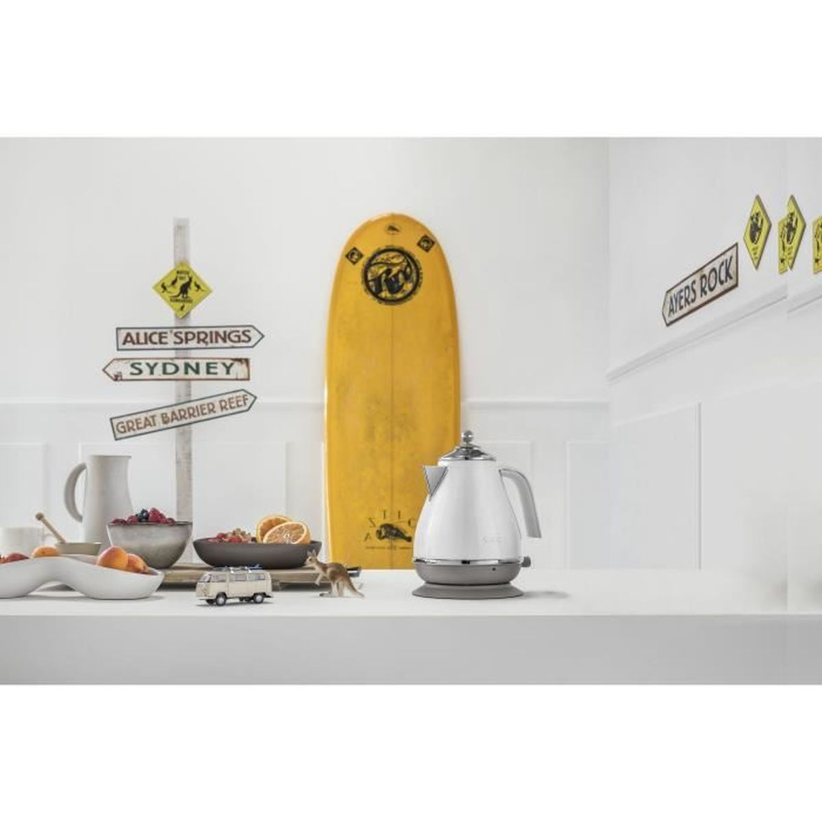 Kitchen scene with a kettle, surfboard, signs, and bowls of fruit. White and yellow dominant.