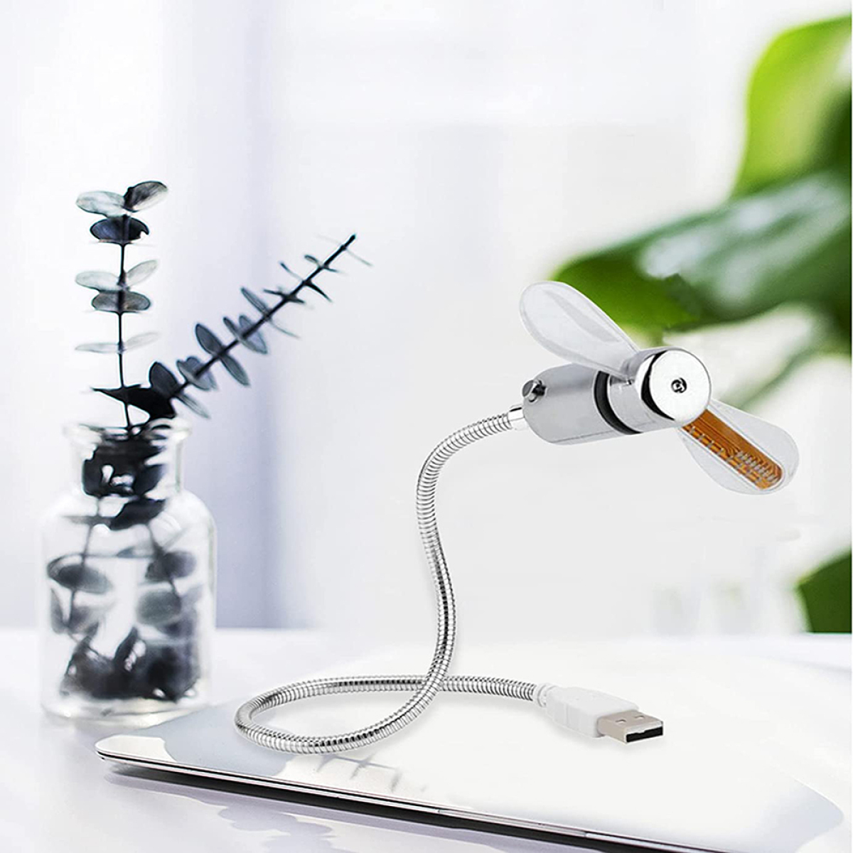 Ein kleiner, flexibler Ventilator mit USB-Anschluss, der auf einer weißen Oberfläche mit einer Vase mit Grünzeug steht.