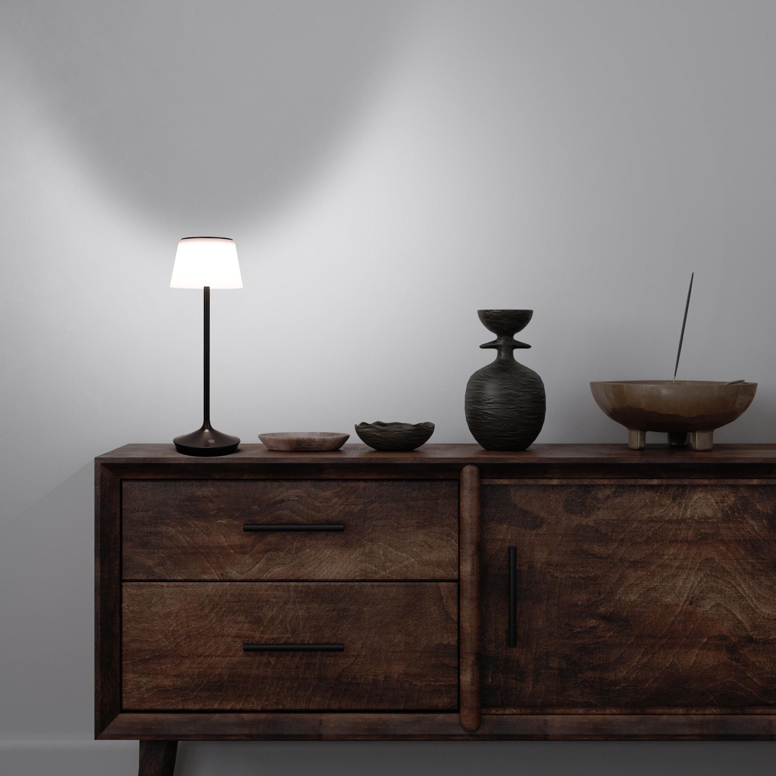 Eine leuchtende Lampe auf einem Holzschrank mit Vasen und Dekorationen. Graue Wand im Hintergrund.