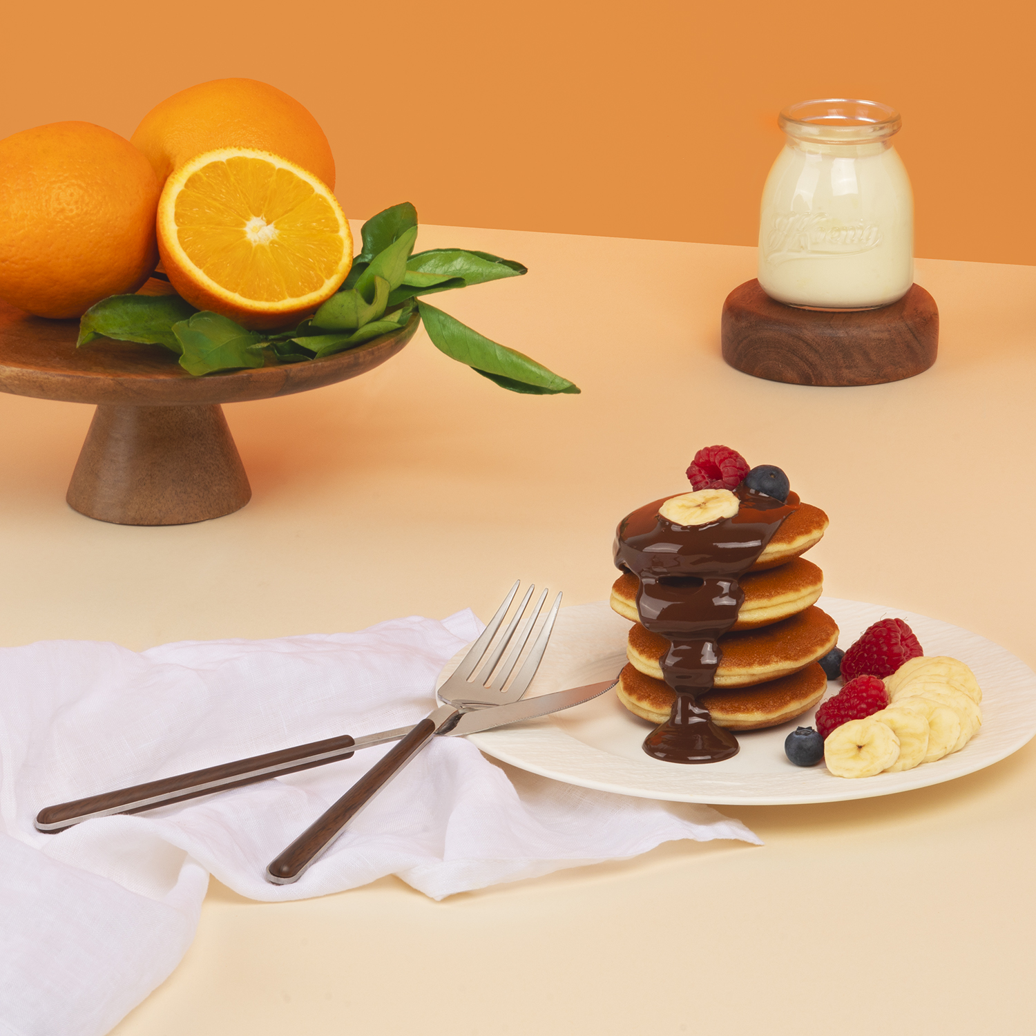 Pancake con salsa al cioccolato, frutti di bosco e banane, arance sullo sfondo.