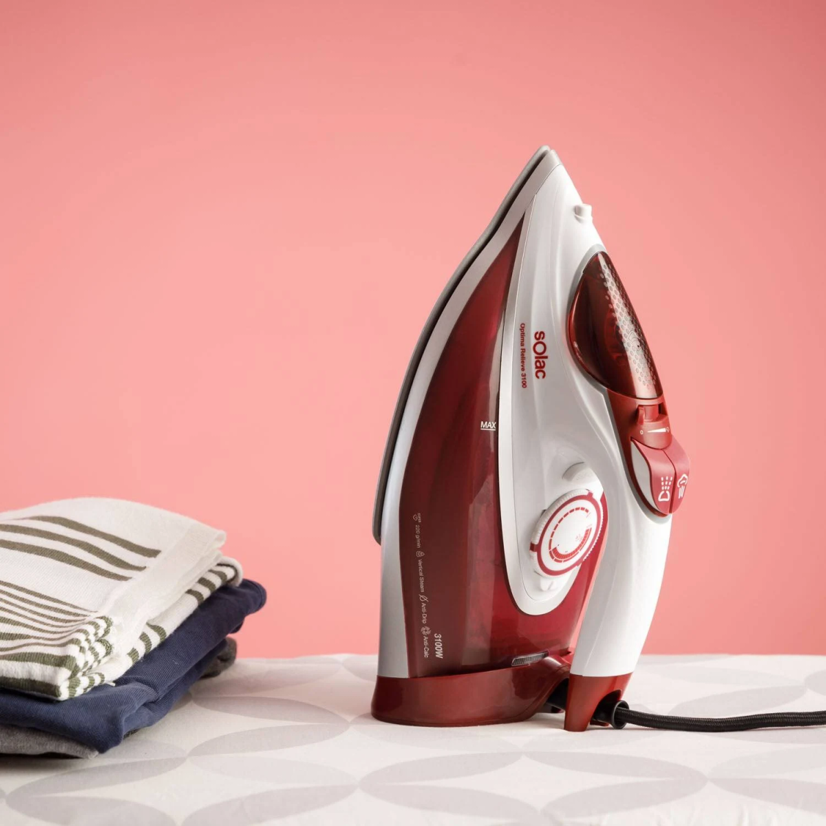 Żelazko na desce z ubraniami. Żelazko jest biało-czerwone. Tło jest różowe.