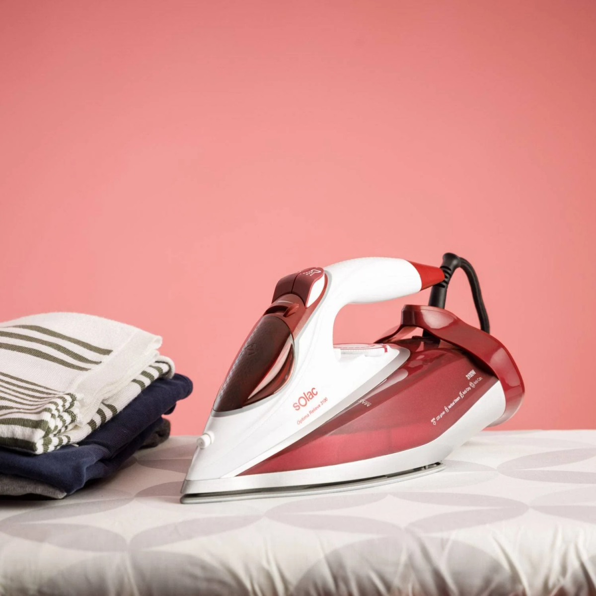 Czerwono-białe żelazko na desce do prasowania, z lewej stos złożonej odzieży.