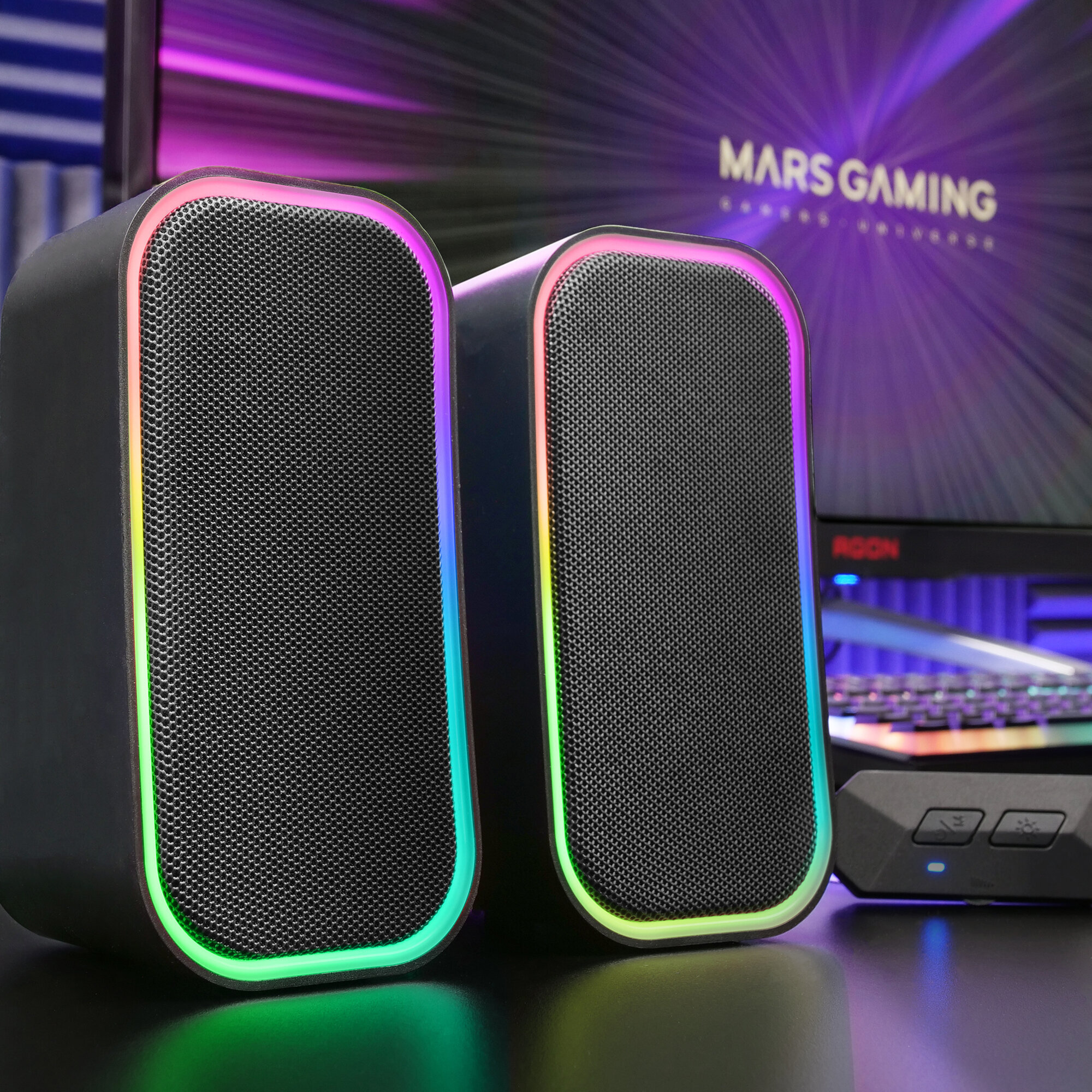 Altavoces negros con luces RGB y panel de control, frente a un monitor.
