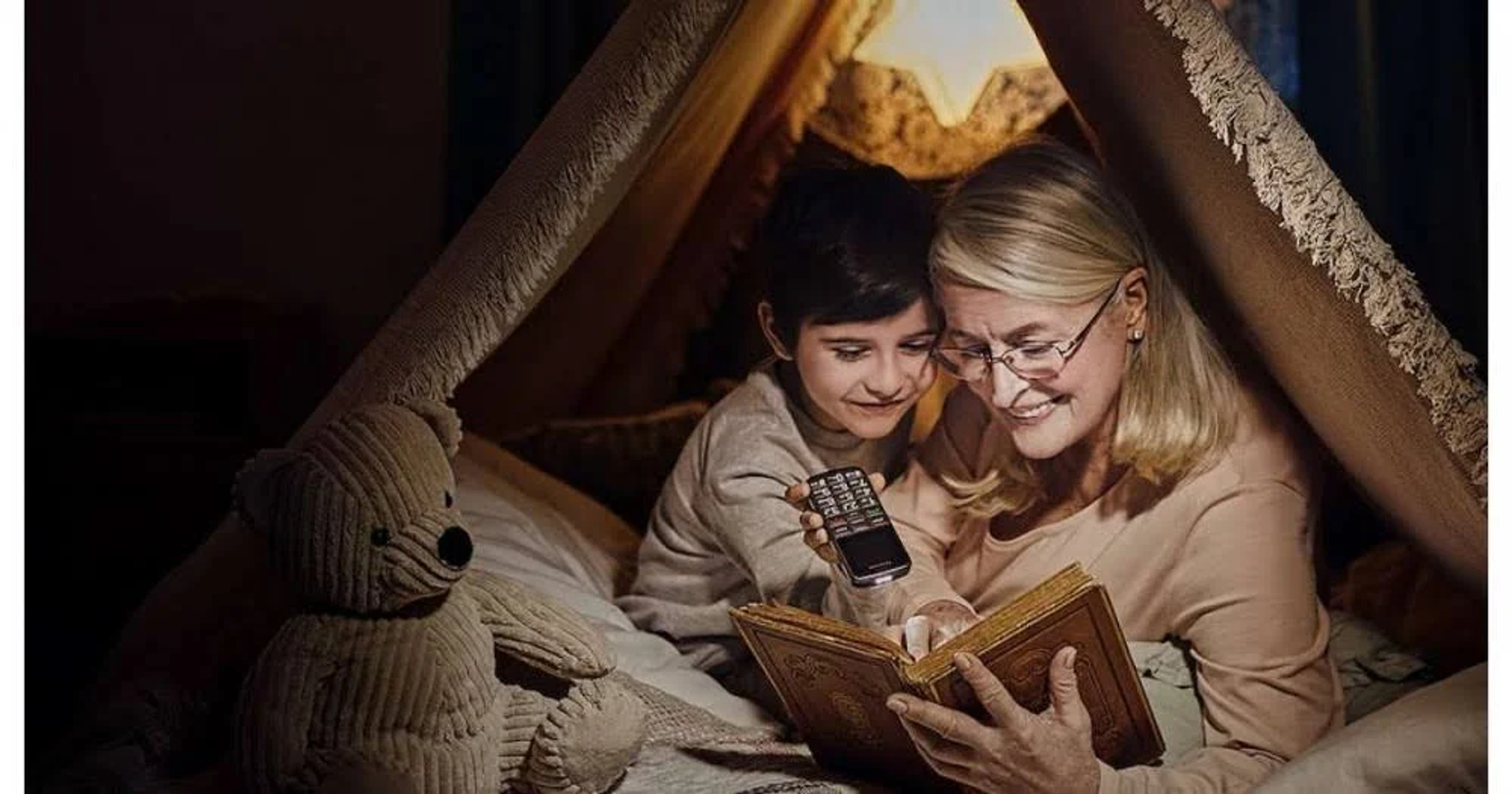 Ein Kind und ein Erwachsener lesen in einem Zelt ein Buch. Ein Teddybär ist in der Nähe.