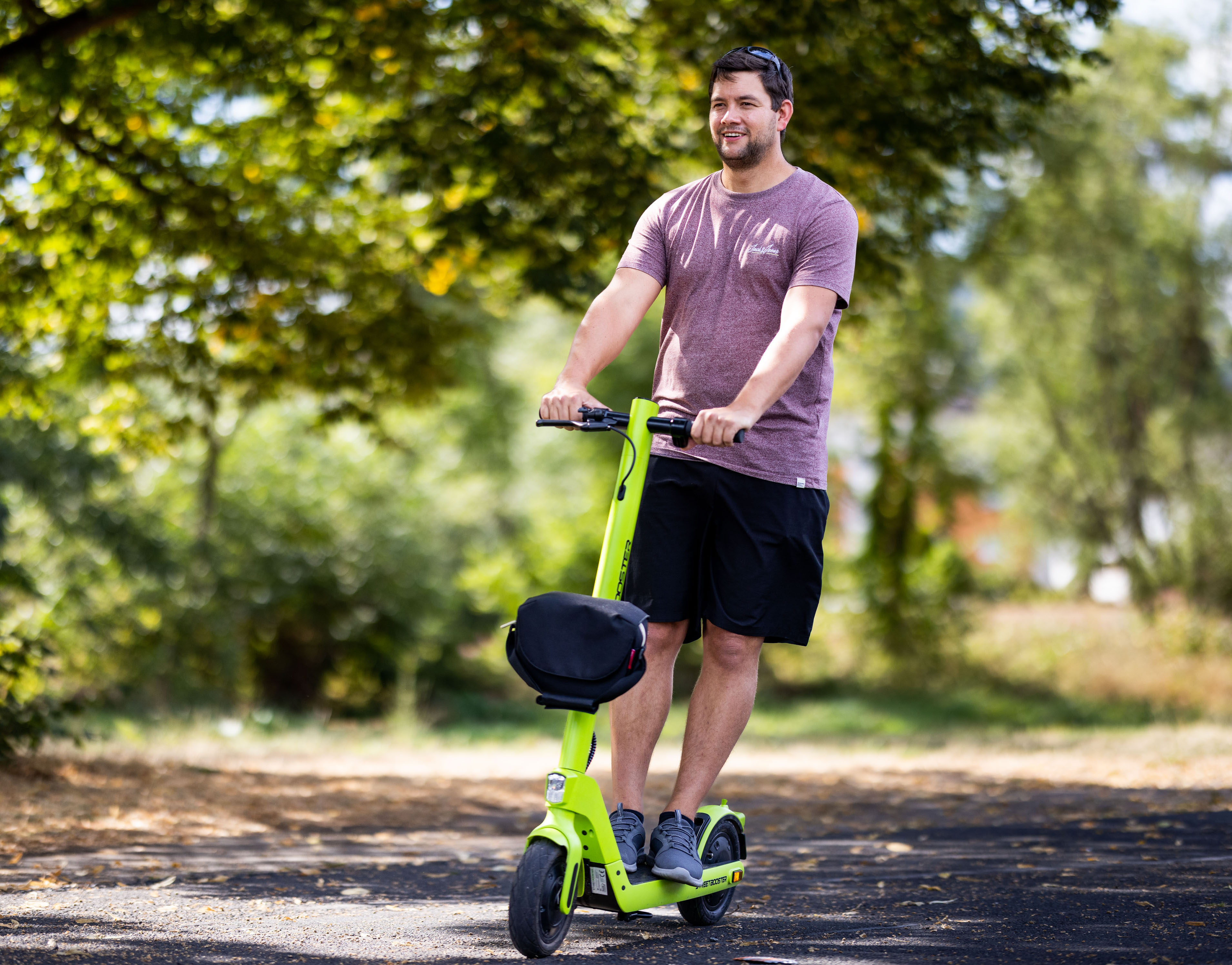 Ein Mann fährt auf einem lindgrünen Roller auf einem Pfad, lächelt, mit verschwommenem Hintergrund.