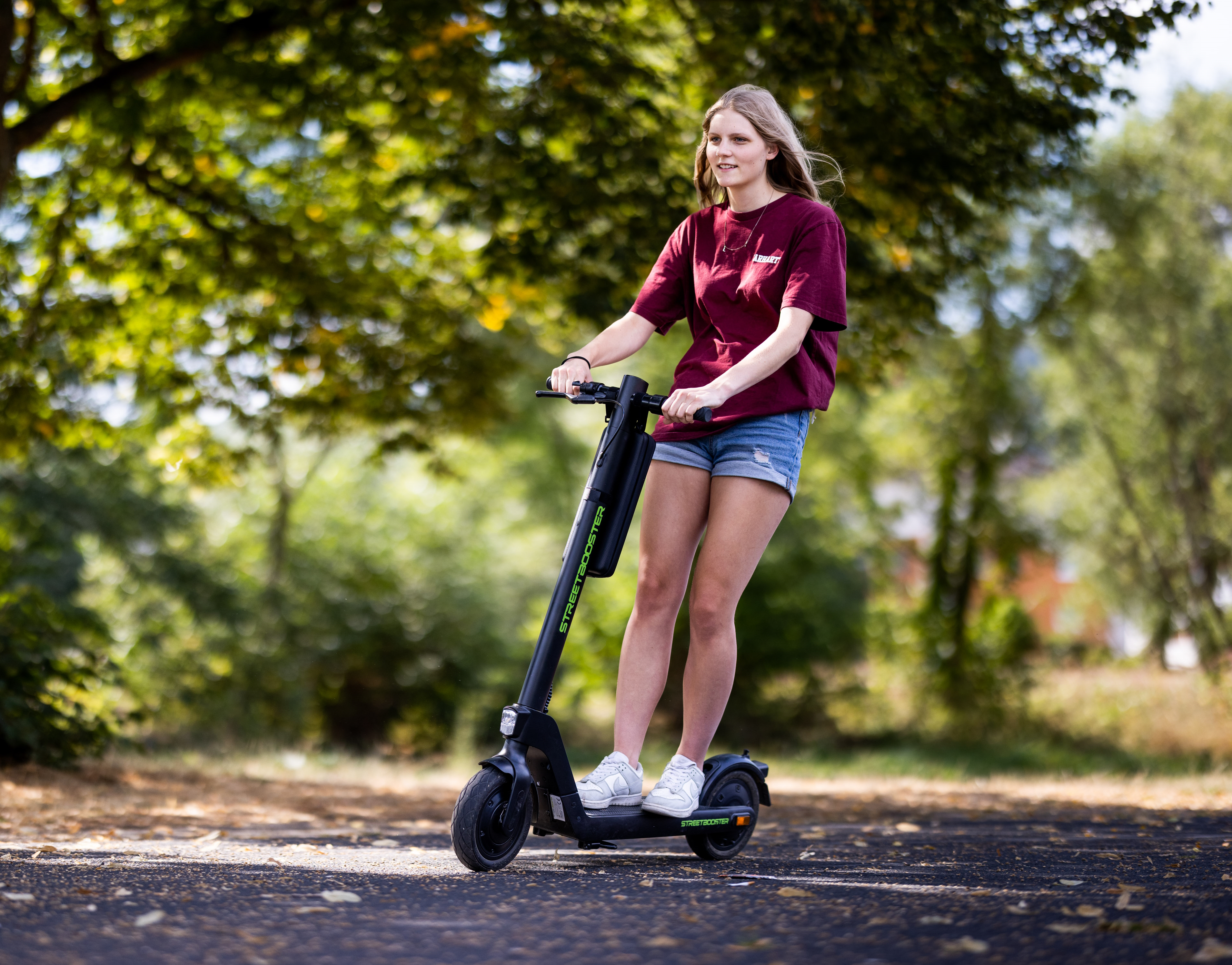 Eine junge Frau fährt mit einem schwarzen Elektroroller auf einem gepflasterten Weg im Park.