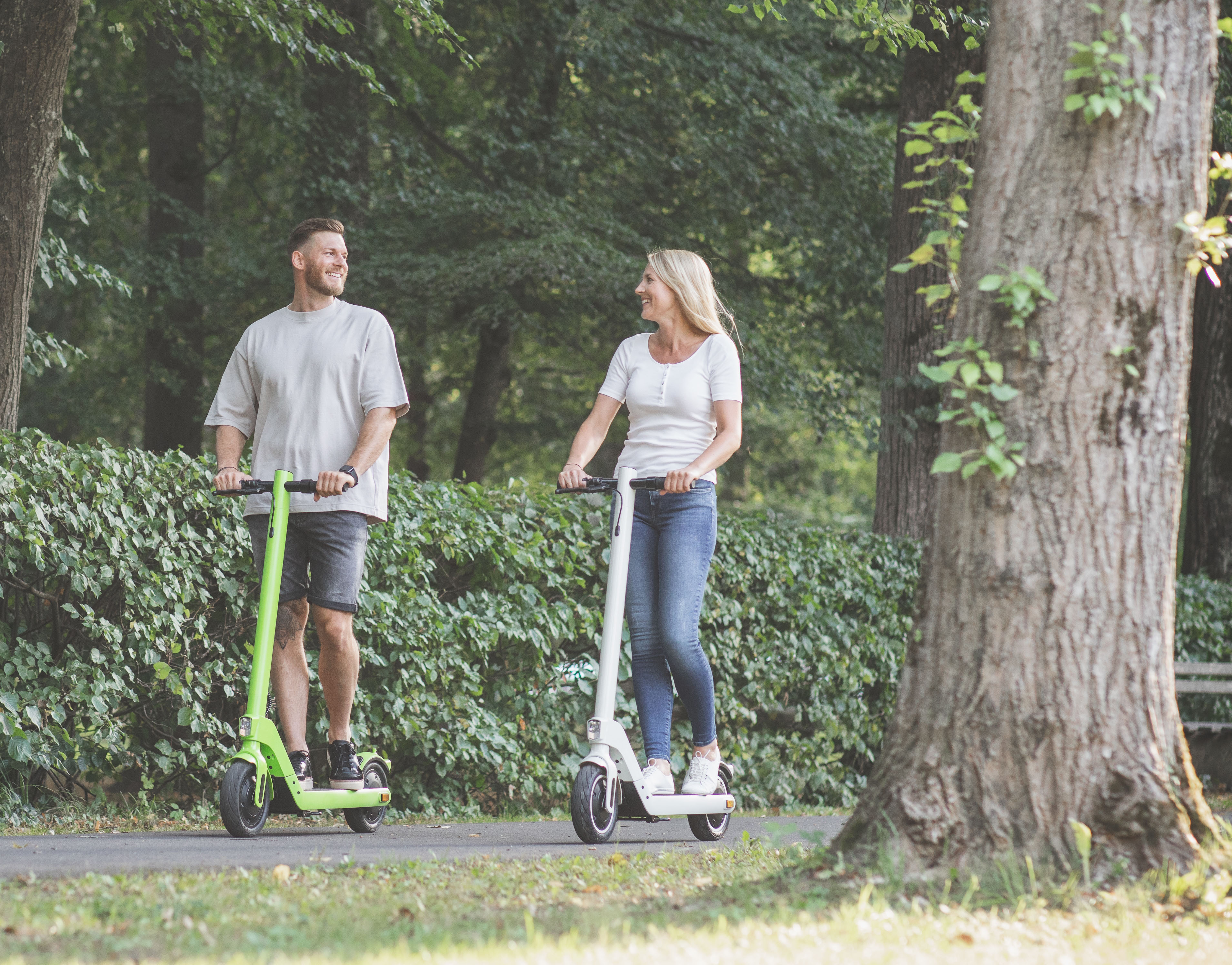 Ein Mann und eine Frau fahren mit Elektrorollern auf einem Weg. Grüne Bäume und Büsche im Hintergrund. Sonniger Tag.