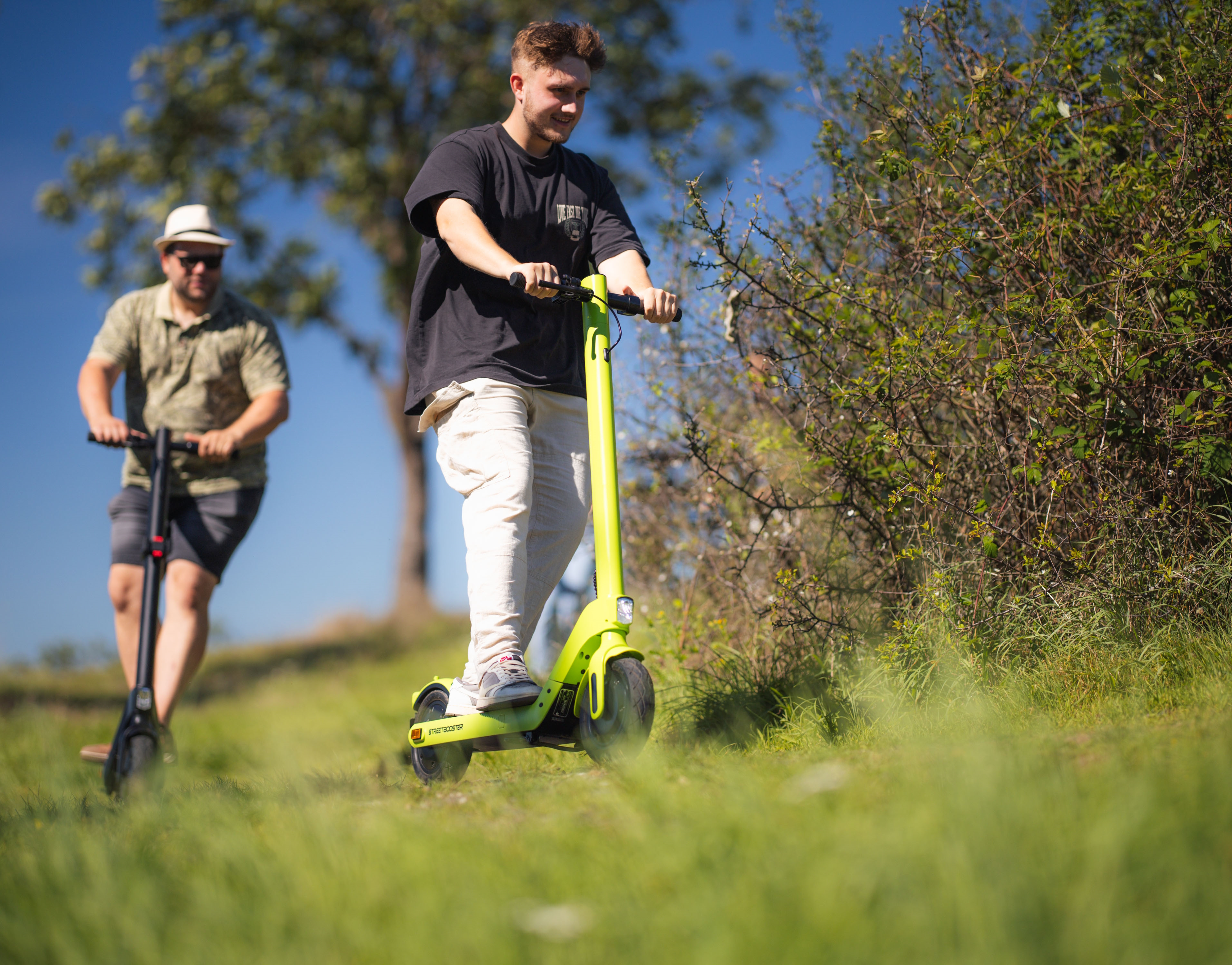 Zwei Männer auf Elektrorollern, einer grün, einer schwarz, auf einem grasbewachsenen Weg mit Bäumen und Büschen.