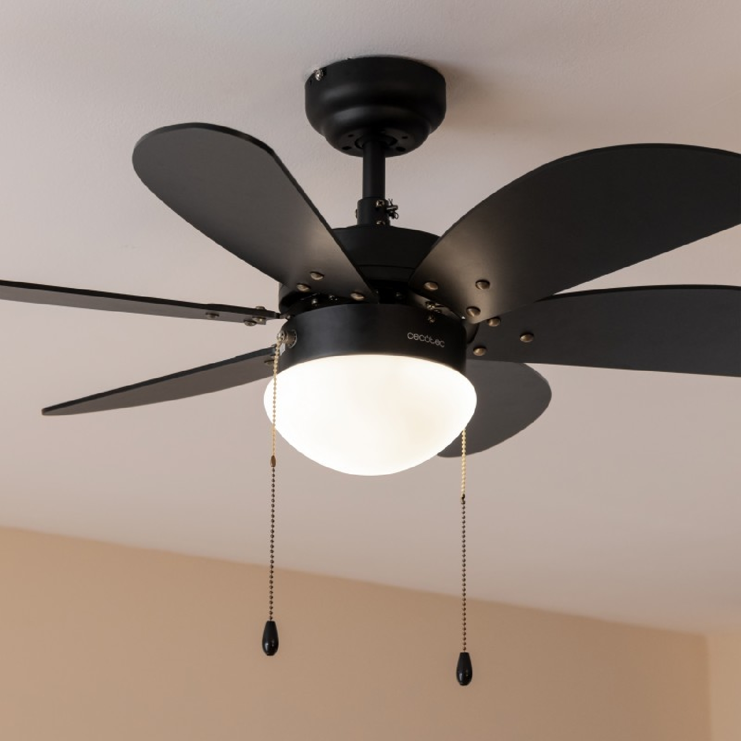 Un ventilatore a soffitto con pale nere e lampada bianca. Soffitto beige e parete beige chiaro.