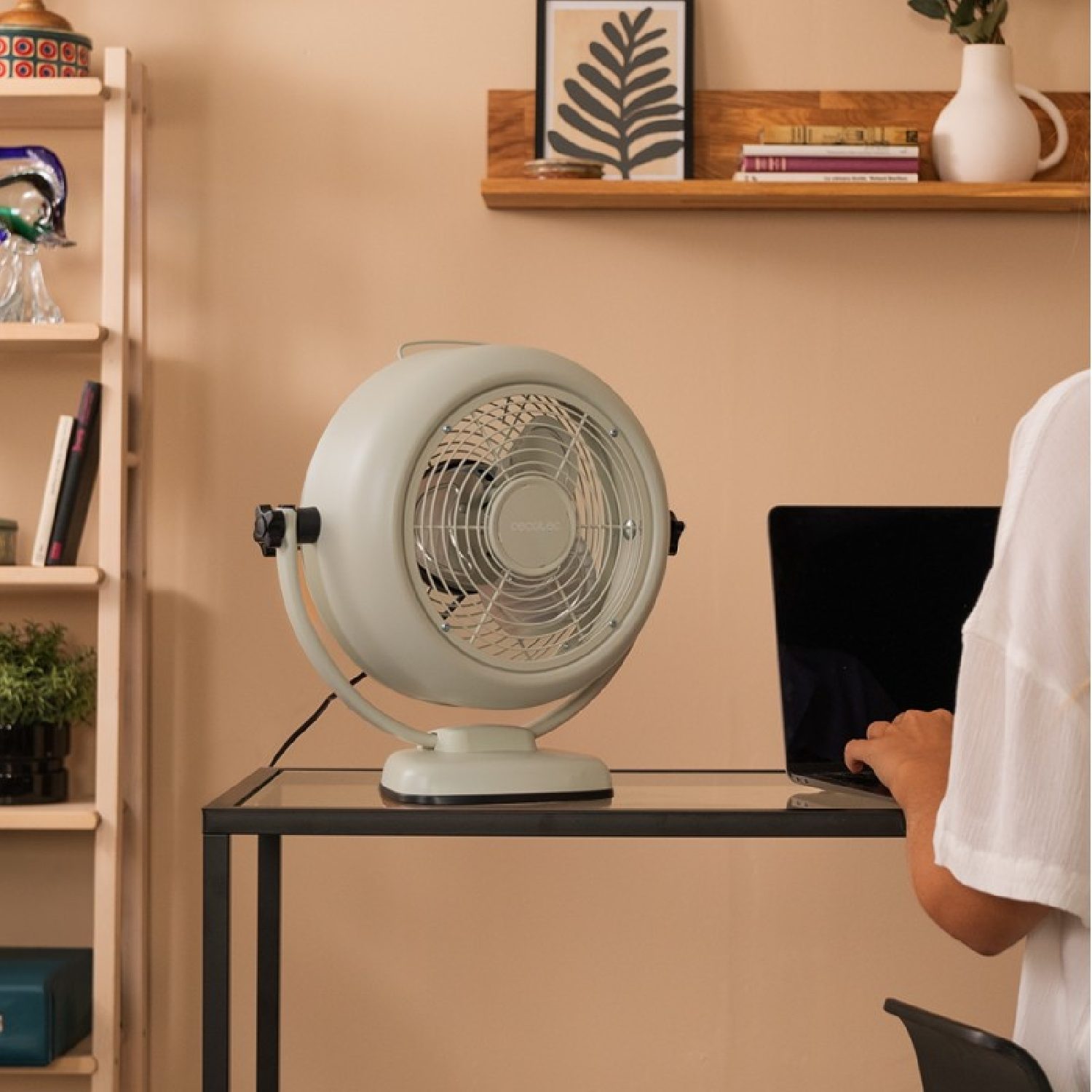 Un ventilatore su una scrivania accanto a un laptop. Libreria sullo sfondo.