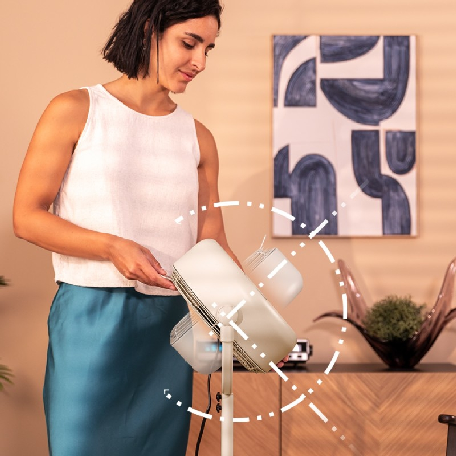 Mujer ajustando un ventilador blanco; pintura y muebles de madera al fondo.