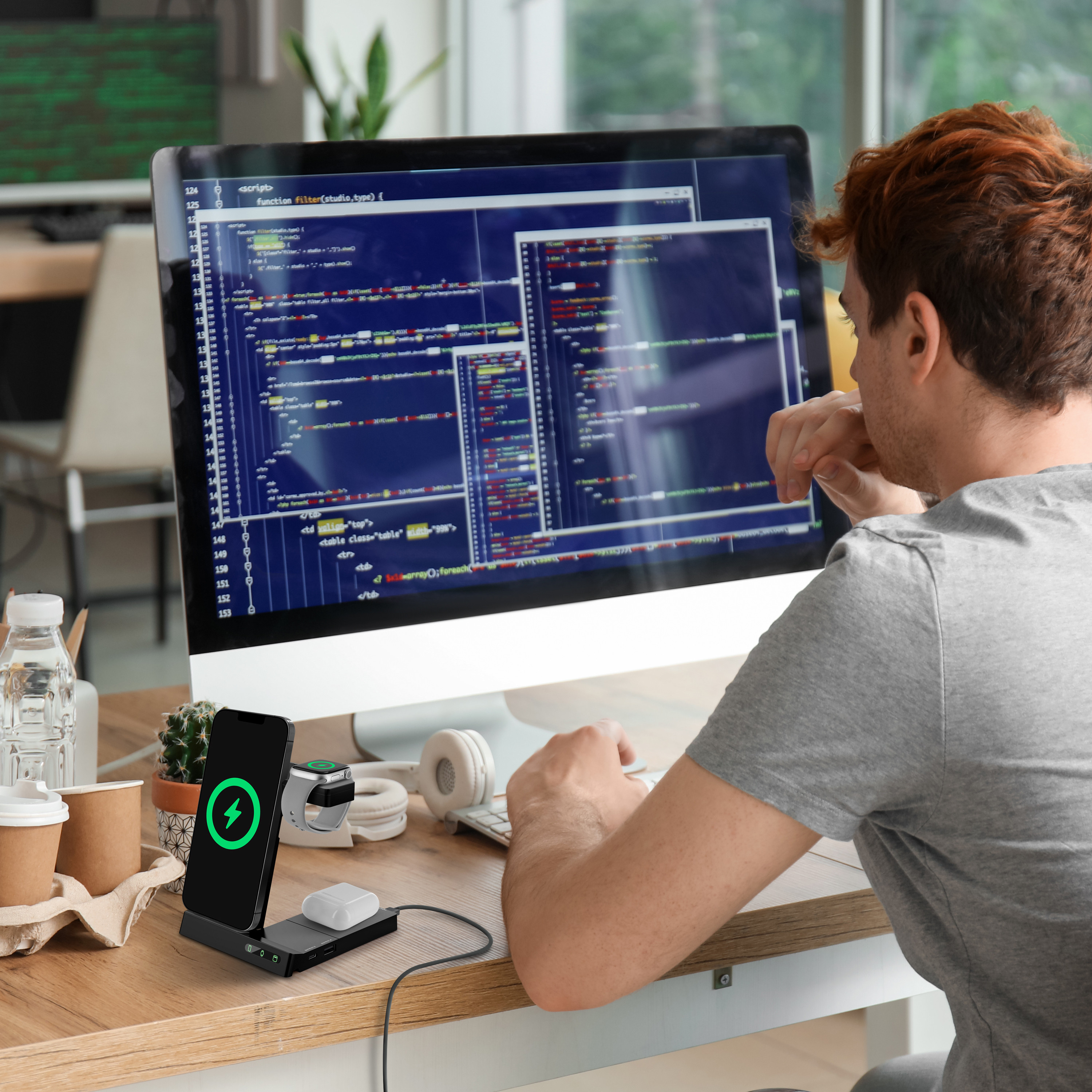 Un hombre en un escritorio con un monitor mostrando código, una estación de carga y un reloj inteligente.