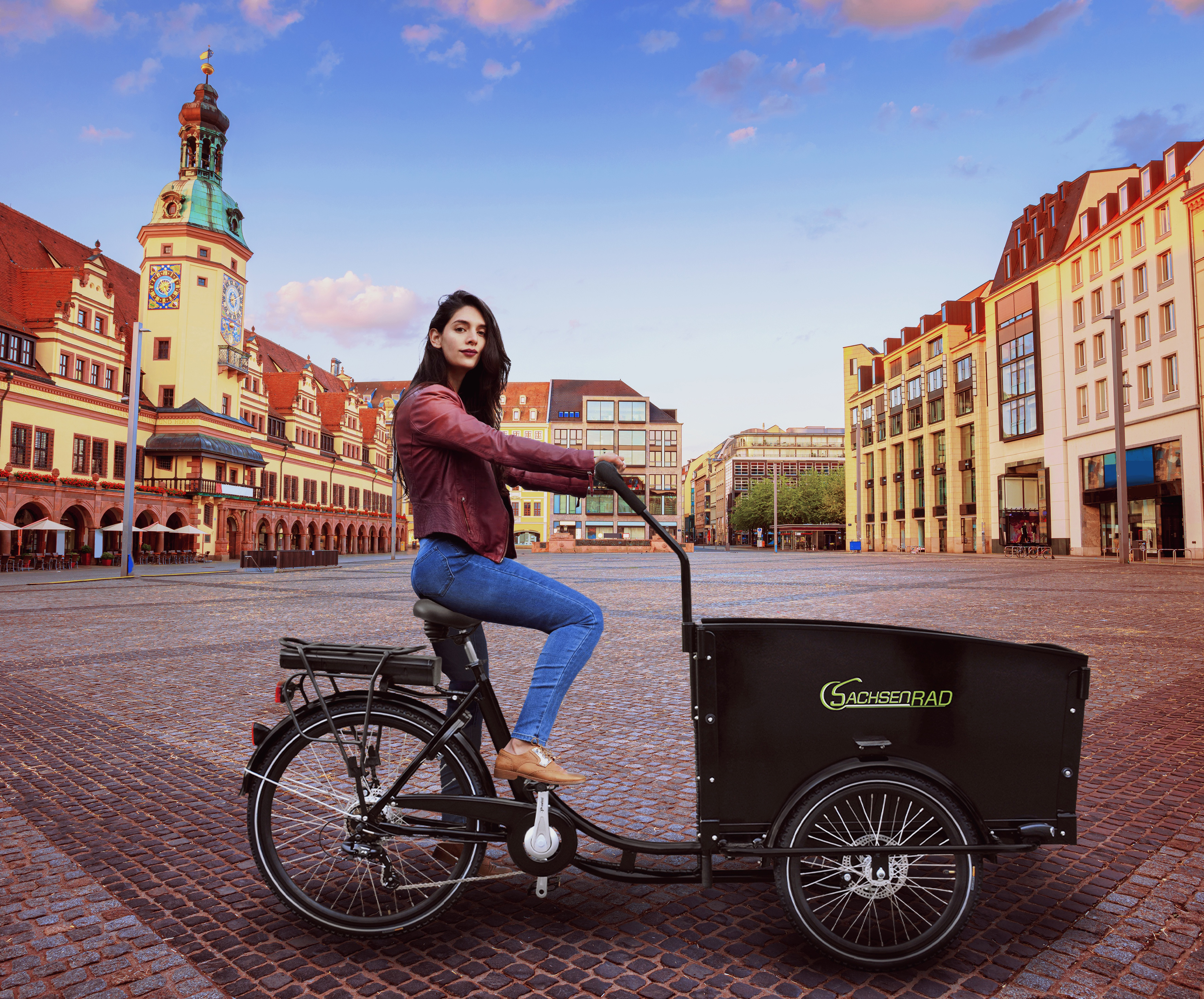 Frau auf einem schwarzen Lastenrad. Stadtplatz im Hintergrund mit Gebäuden und blauem Himmel.
