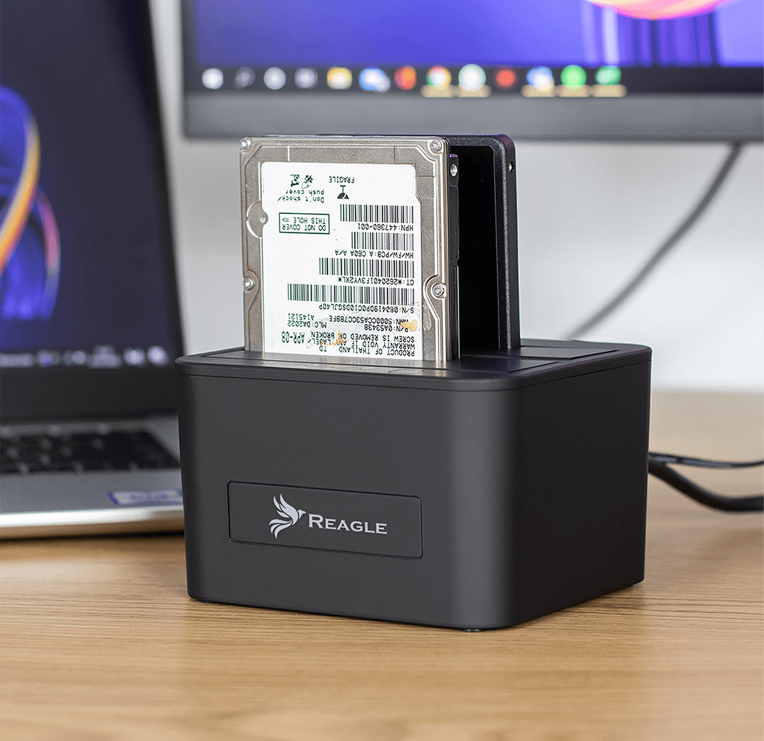 Czarna stacja dokująca dysku twardego z dyskiem twardym w środku, na drewnianym biurku, z monitorem komputera.