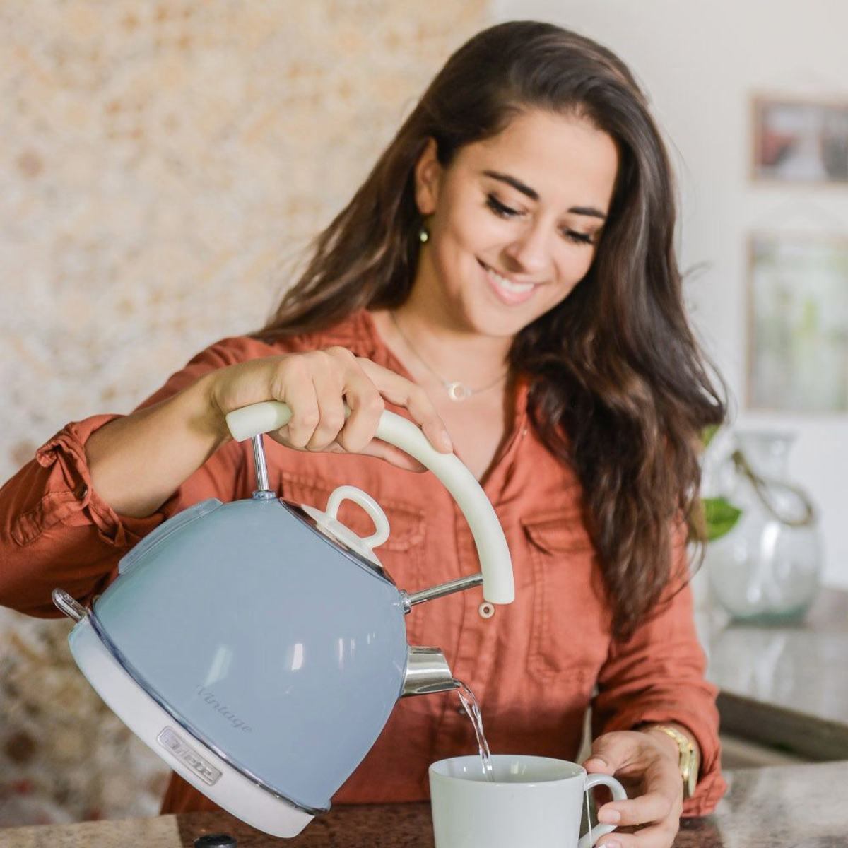 Vrouw giet water uit een blauwe waterkoker in een witte mok; keukenachtergrond.