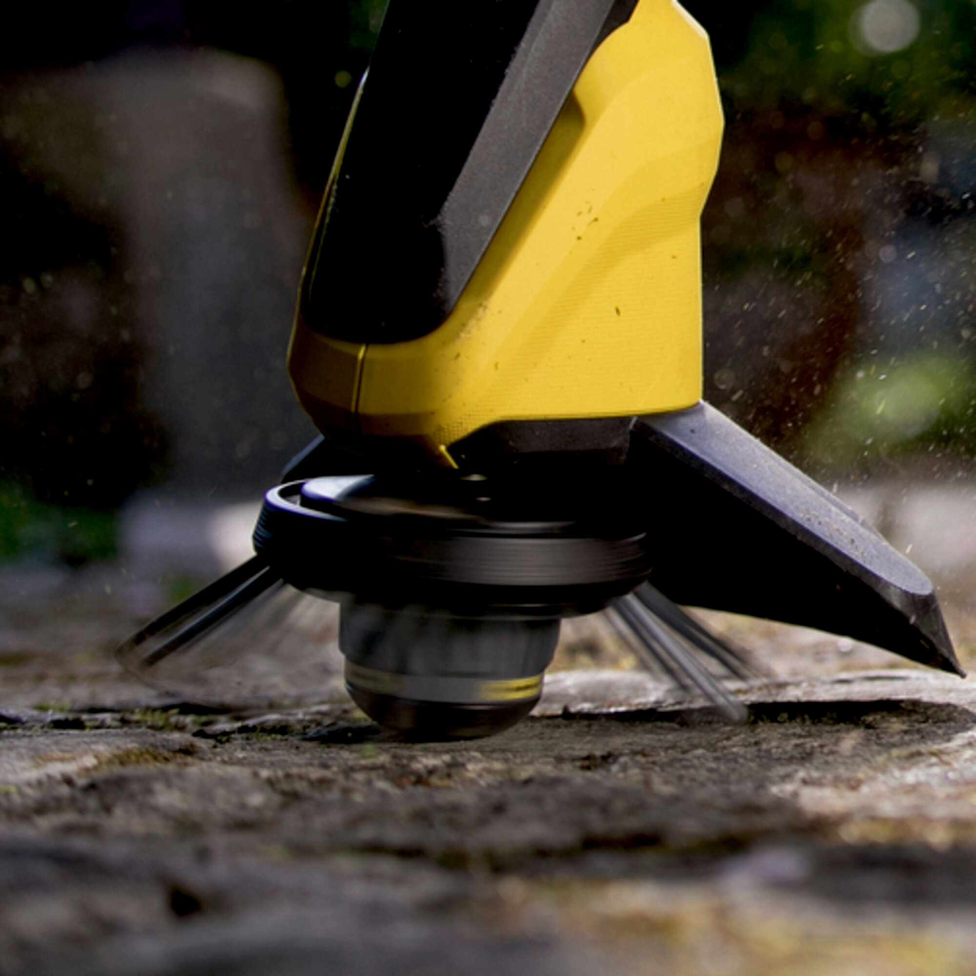 Żółto-czarna głowica podkaszarki, wirująca nad kamieniem, rozmyte tło z zielenią.