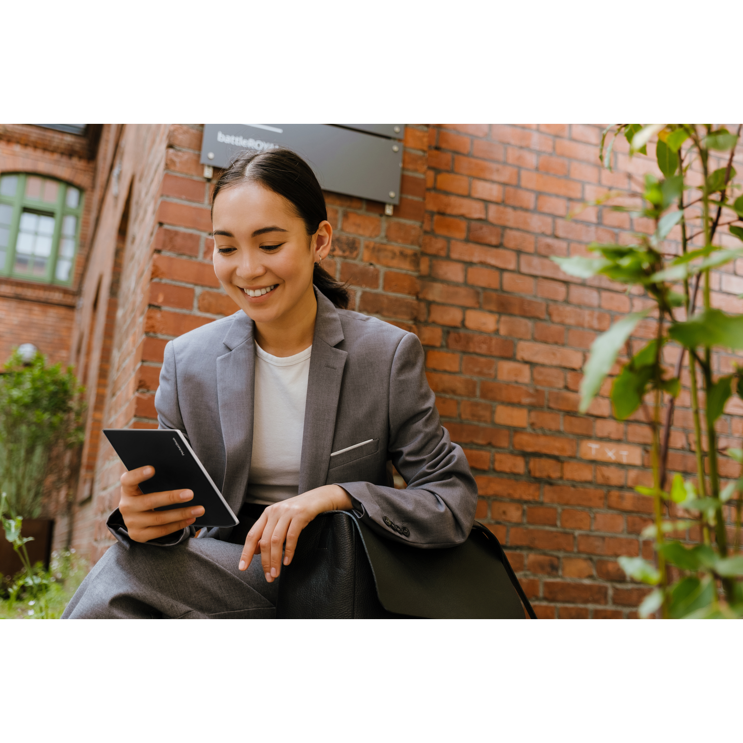 Uśmiechnięta kobieta w szarym garniturze czyta tablet, siedząc pod ceglanym murem z zielonymi roślinami.